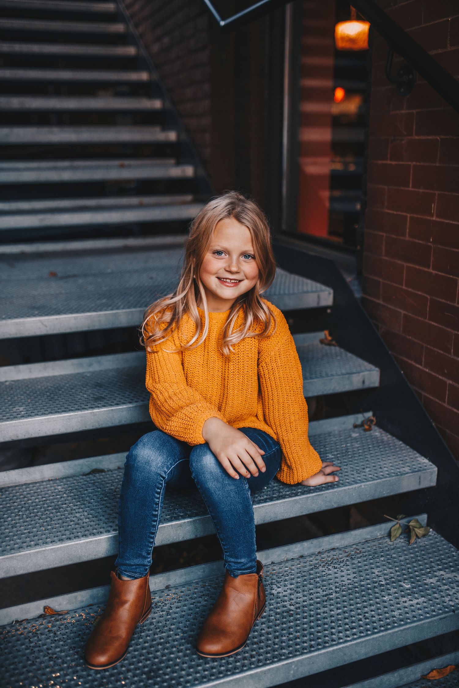 Adorable young girl in mustard yellow knit sweater and jeans sitting on metal stairs, smiling brightly.
