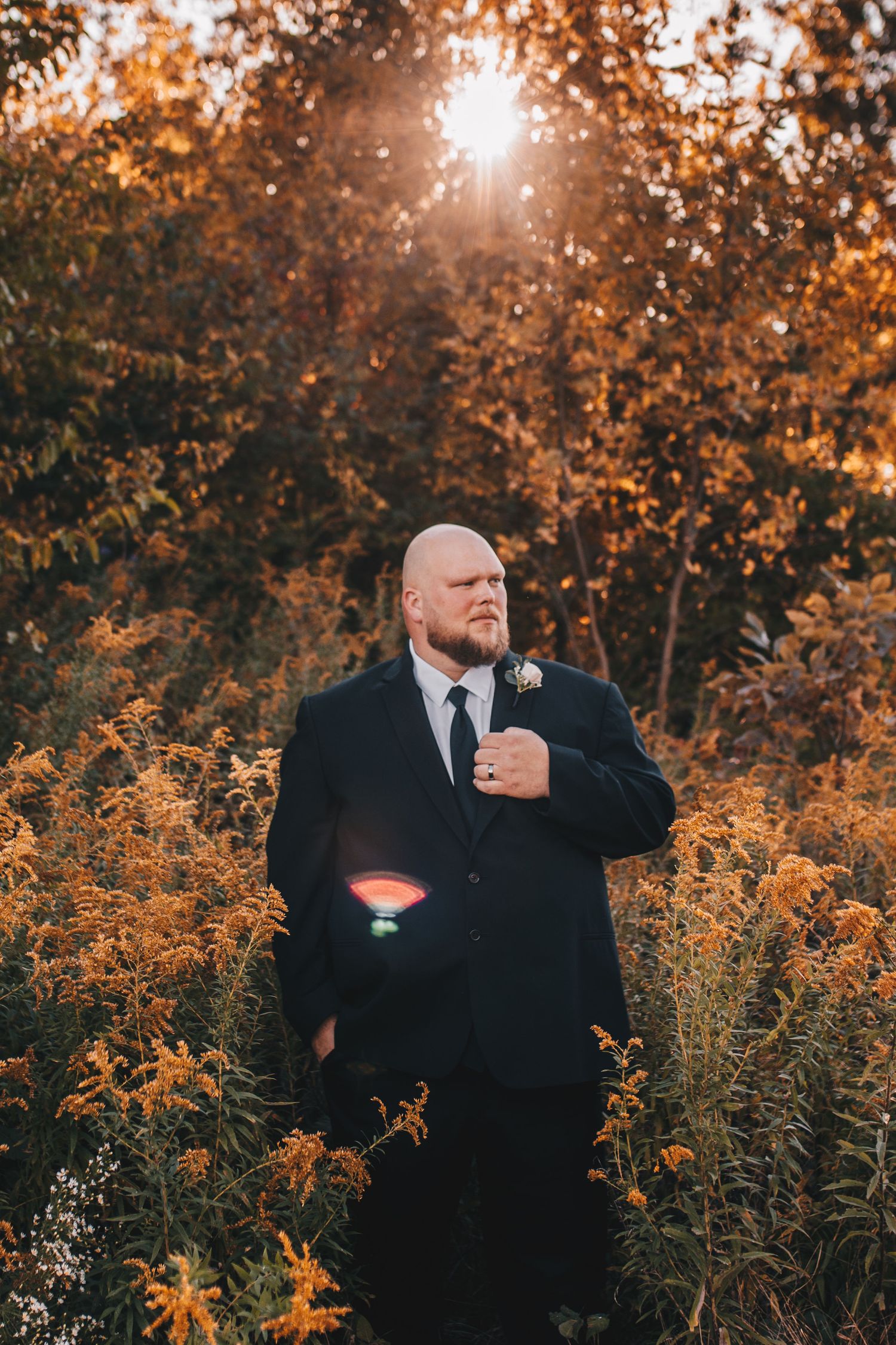 Bald groom in black suit standing in golden autumn wildflower field with sunburst.