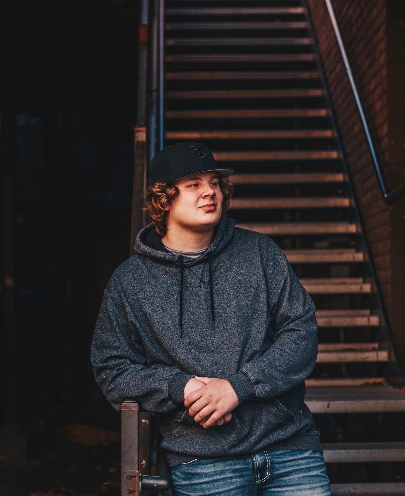 Young man in gray hoodie and black cap leaning against railing near brick staircase.