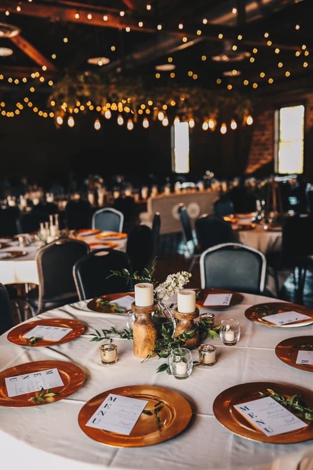 Elegant wedding reception with string lights, copper charger plates, and greenery centerpieces.