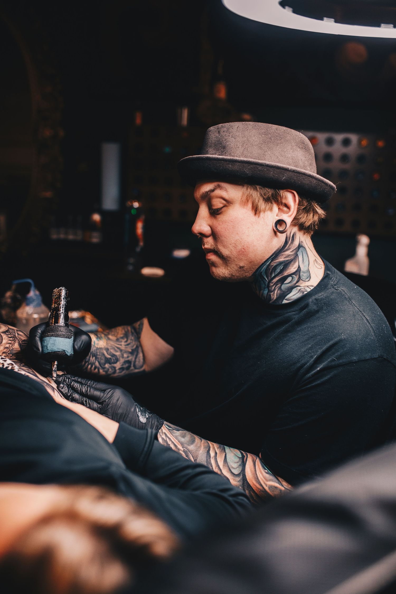 Heavily tattooed artist wearing a brown hat working on a tattoo in a dark studio.