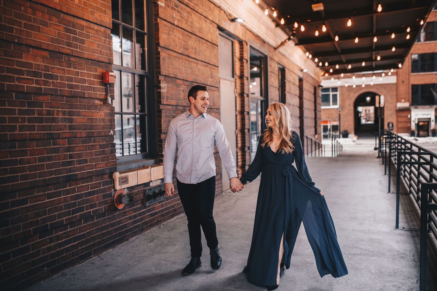 Couple holds hands walking along brick building corridor decorated with string lights during engagement session.