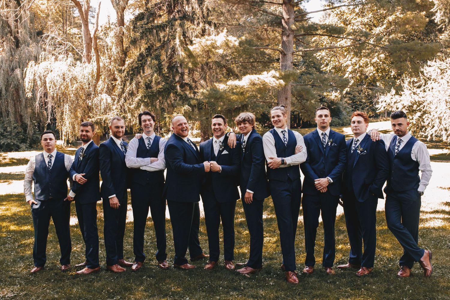 Groom and groomsmen in navy suits posing casually together outdoors under lush green trees.