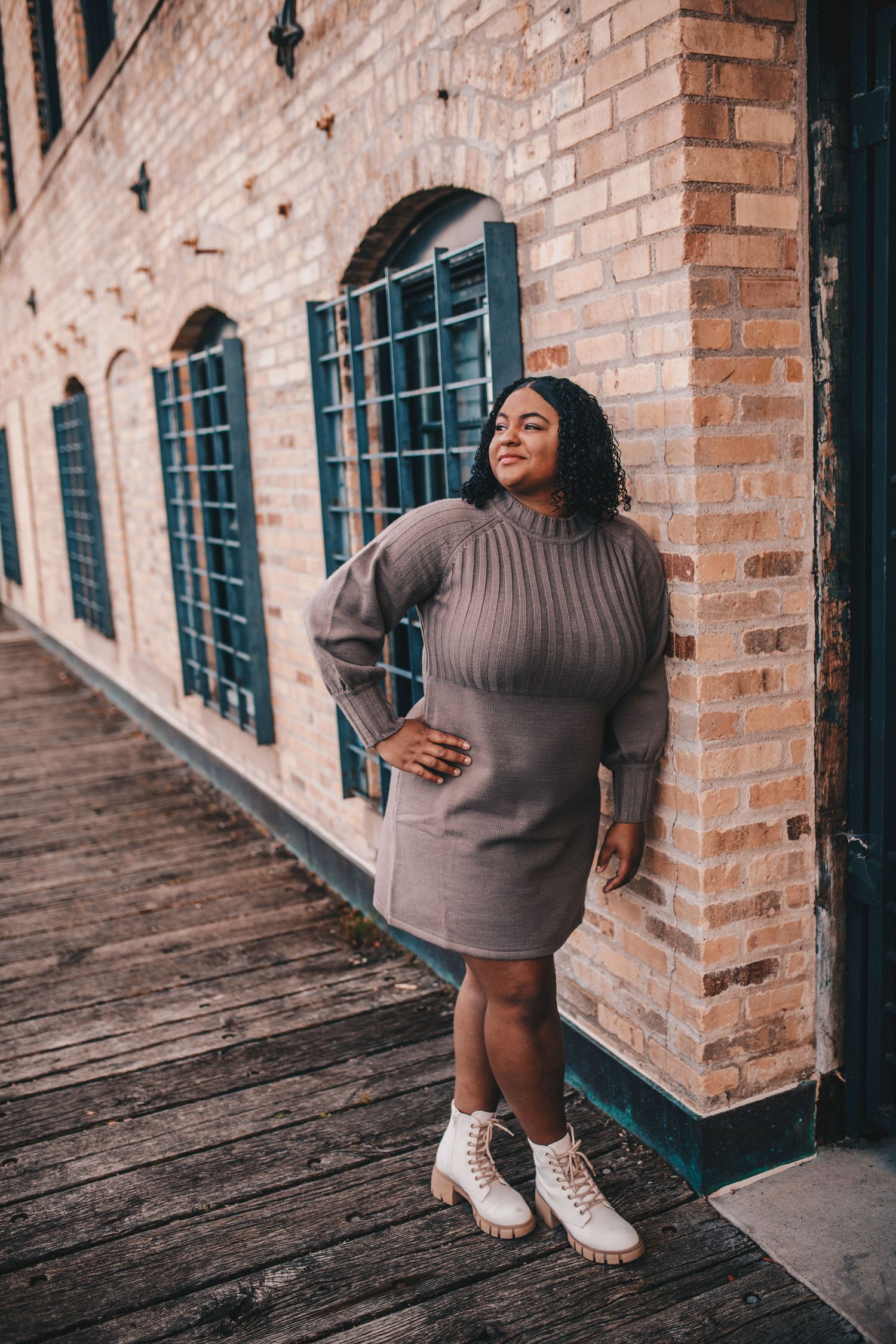 Woman in gray sweater dress and white boots posing by brick building exterior.