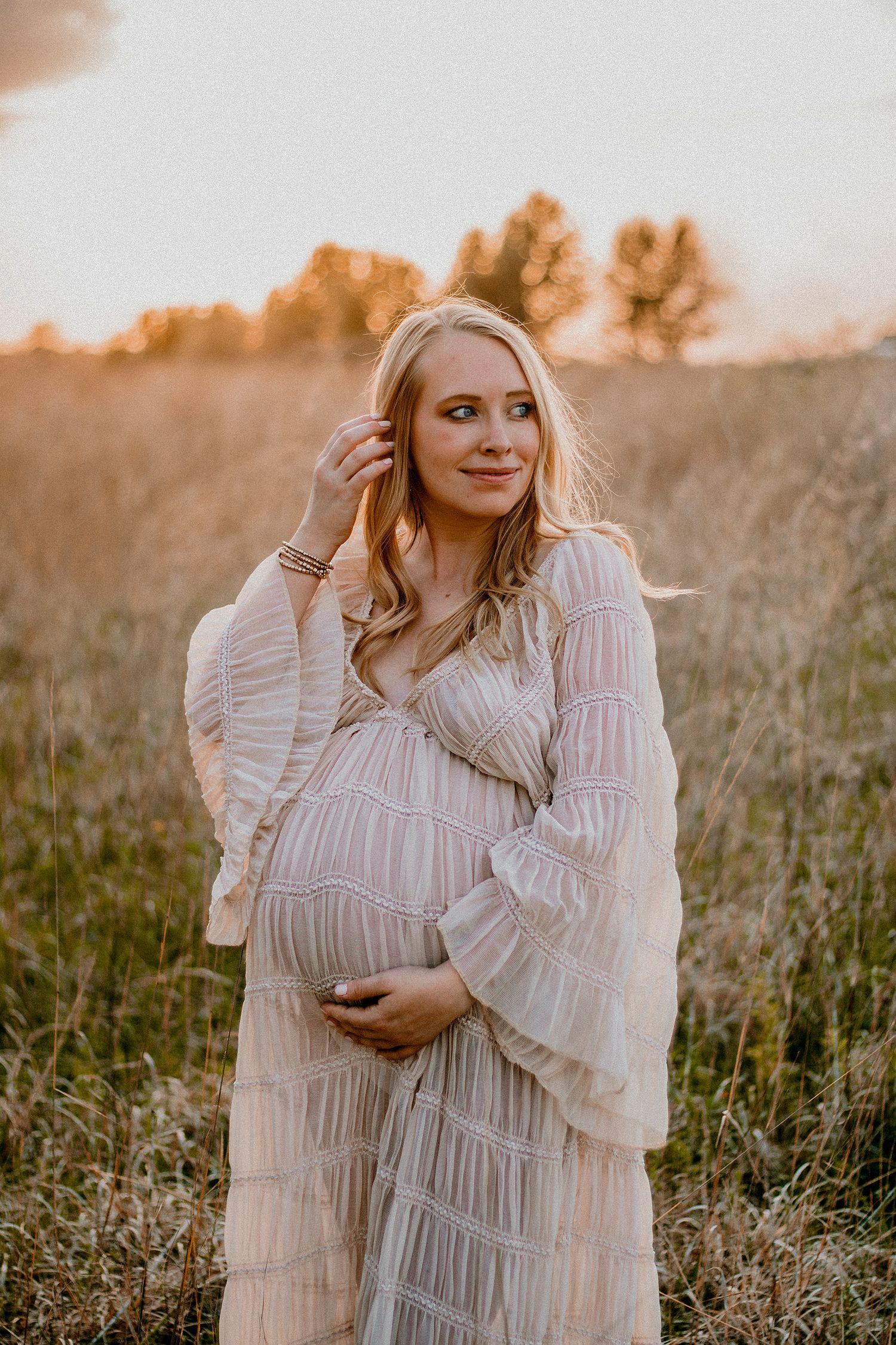 Blonde pregnant woman in boho white dress cradling belly in sunset field.