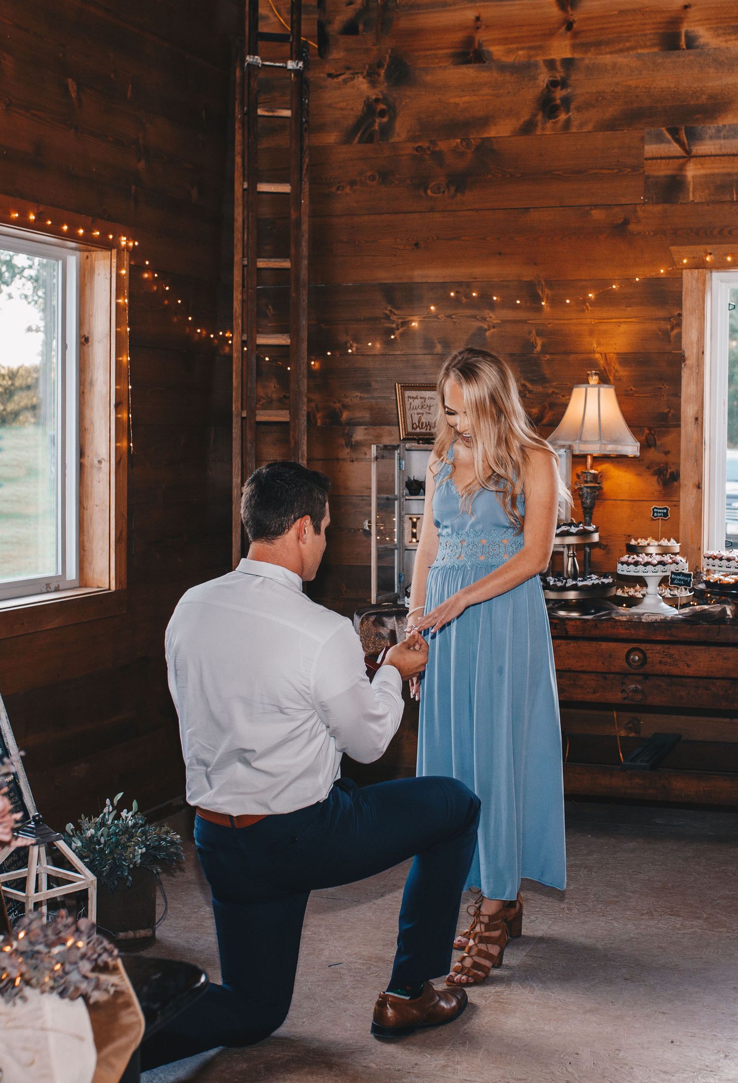 Man proposes on one knee to woman in blue dress inside rustic wooden barn with string lights.