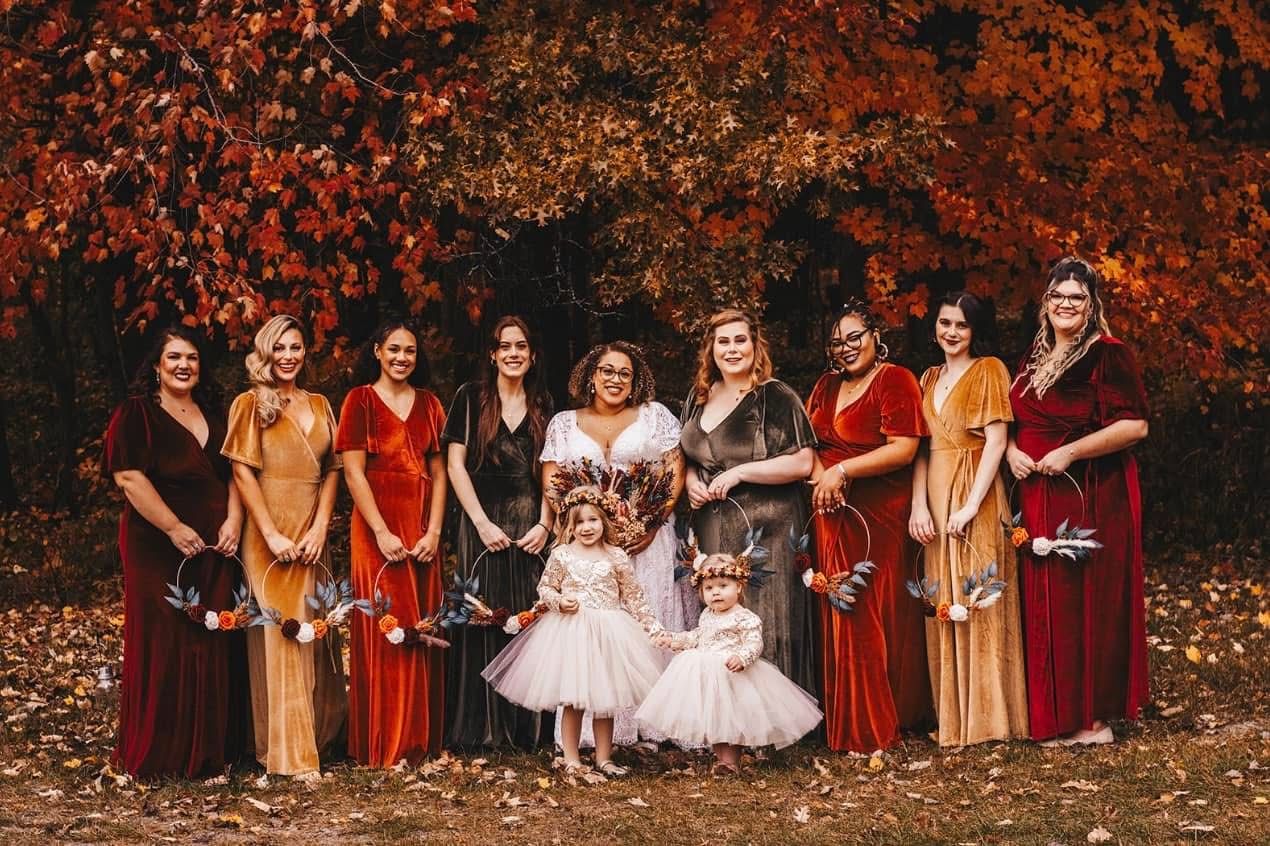 Bridal party in autumn tones with flower girls posing before vibrant red fall foliage.
