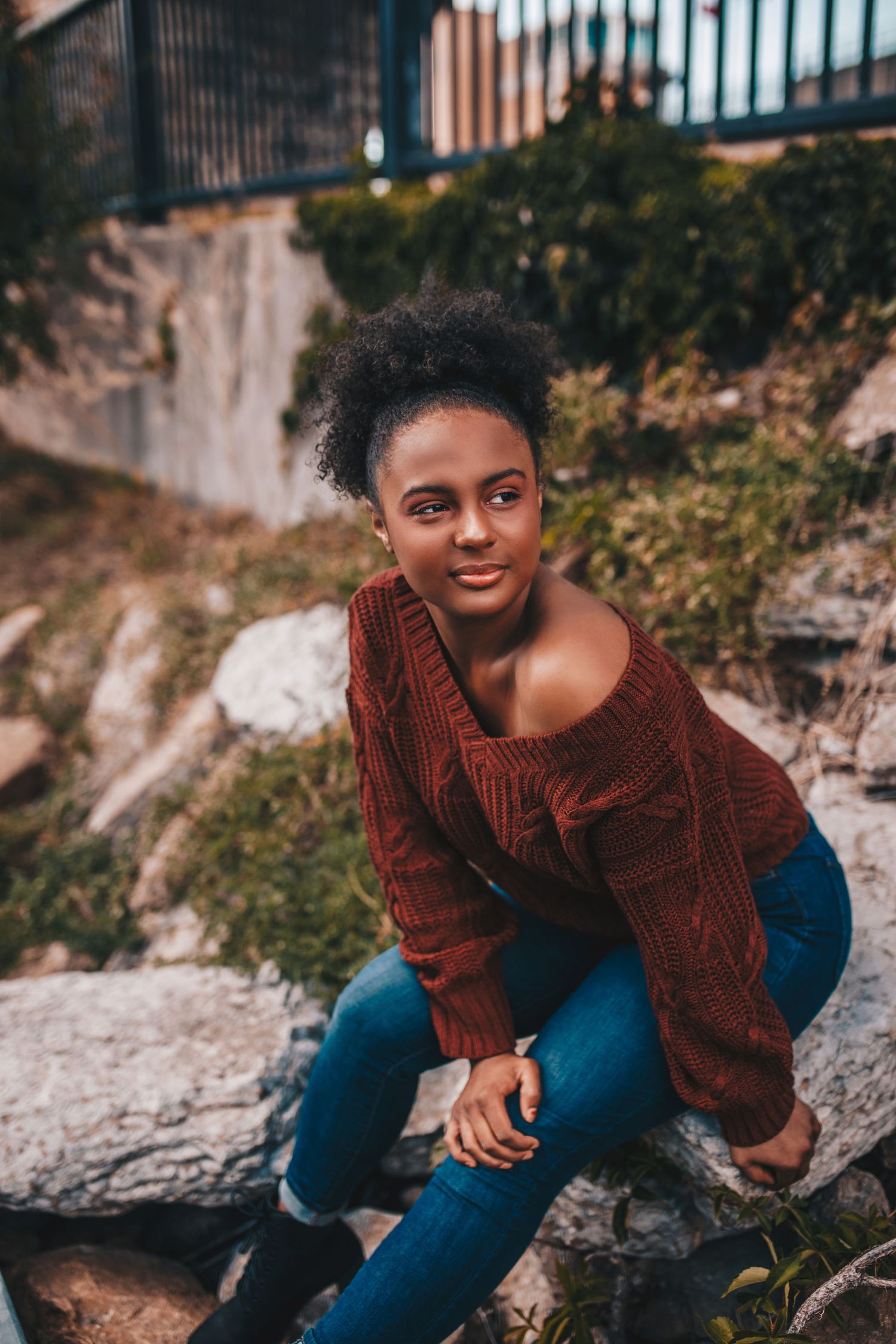 Young woman in red sweater and jeans sitting on rocks near waterfall.