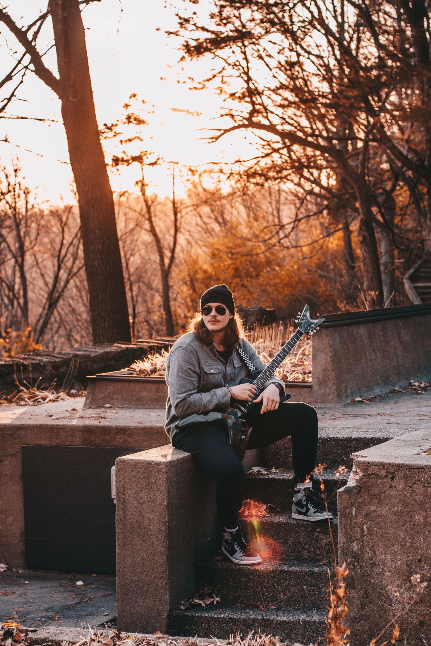 Young woman with red hair sits on stone steps holding electric guitar at golden hour in autumn park.