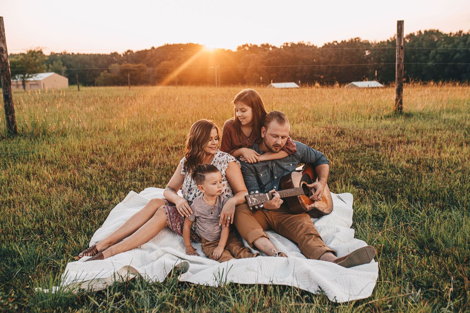 Family of four sitting on blanket in golden field at sunset, father playing guitar while children and mother listen.
