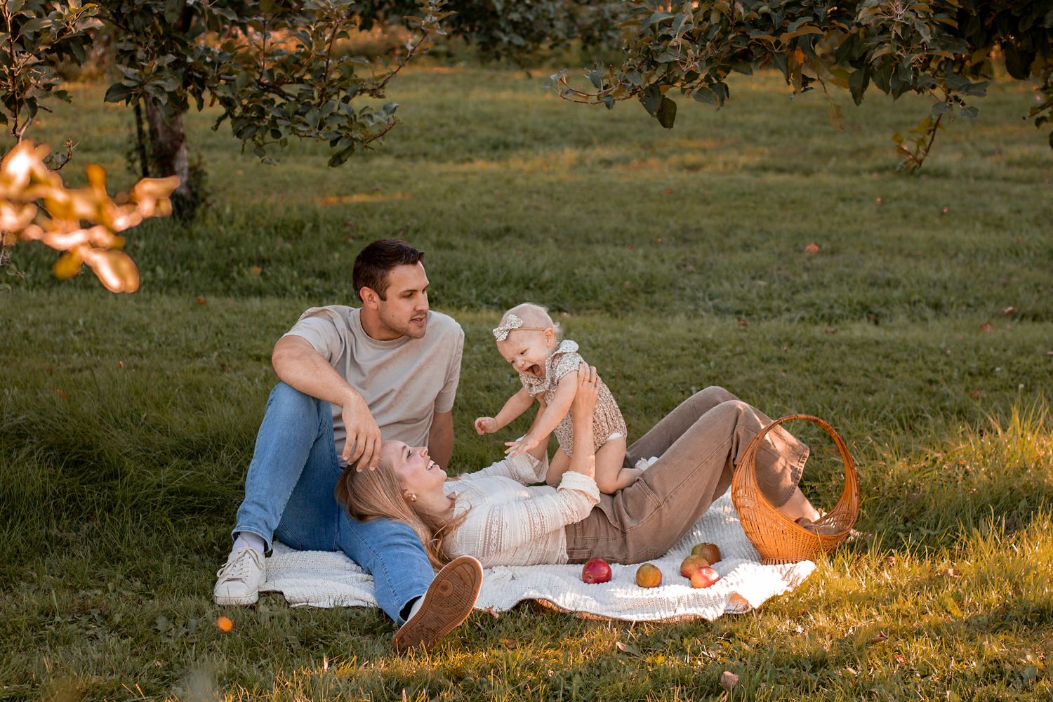 Family enjoying picnic on grass with baby in sunny orchard setting.