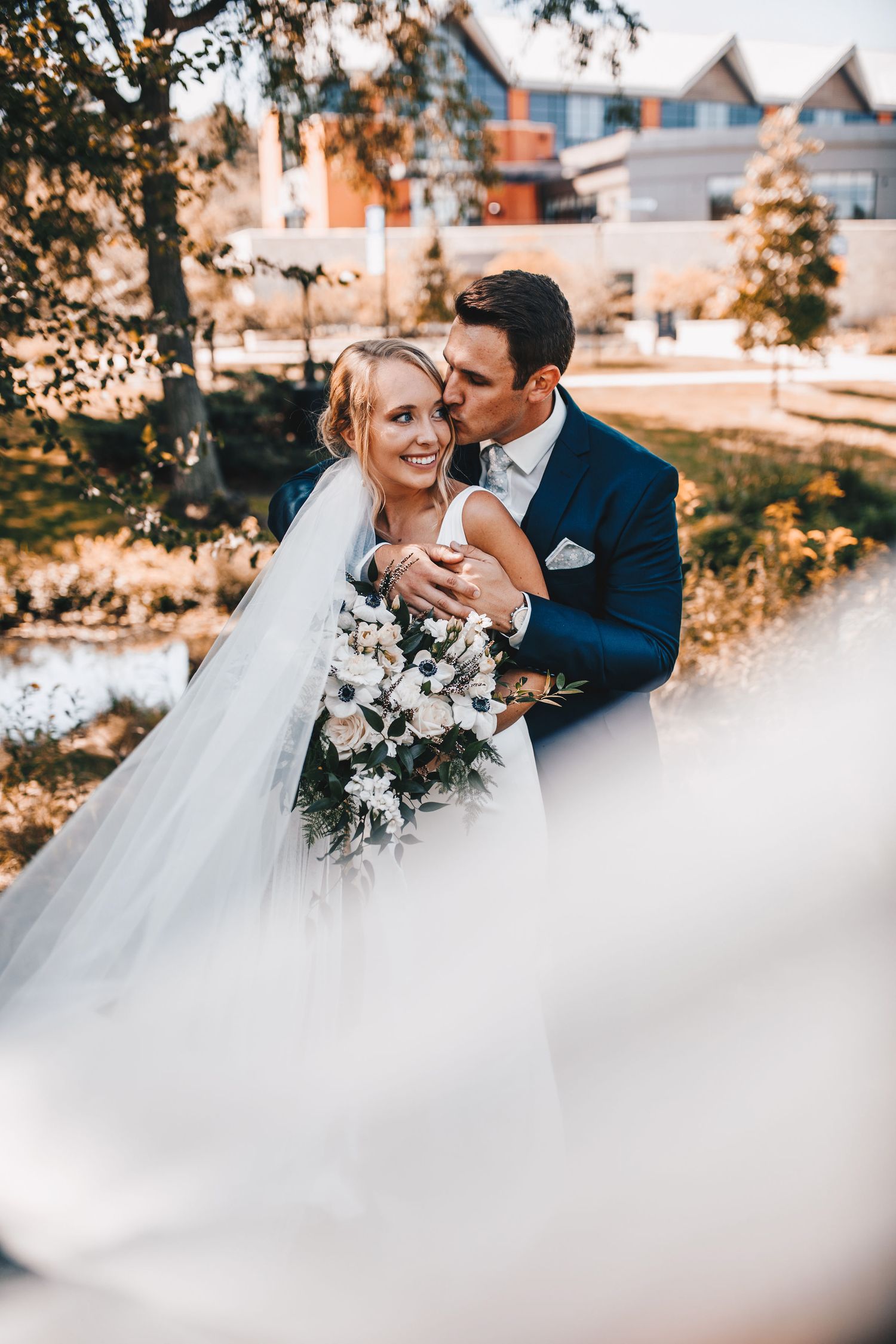 Bride and groom embrace outdoors, she holds white floral bouquet, veil flowing, groom in navy suit kisses her cheek.