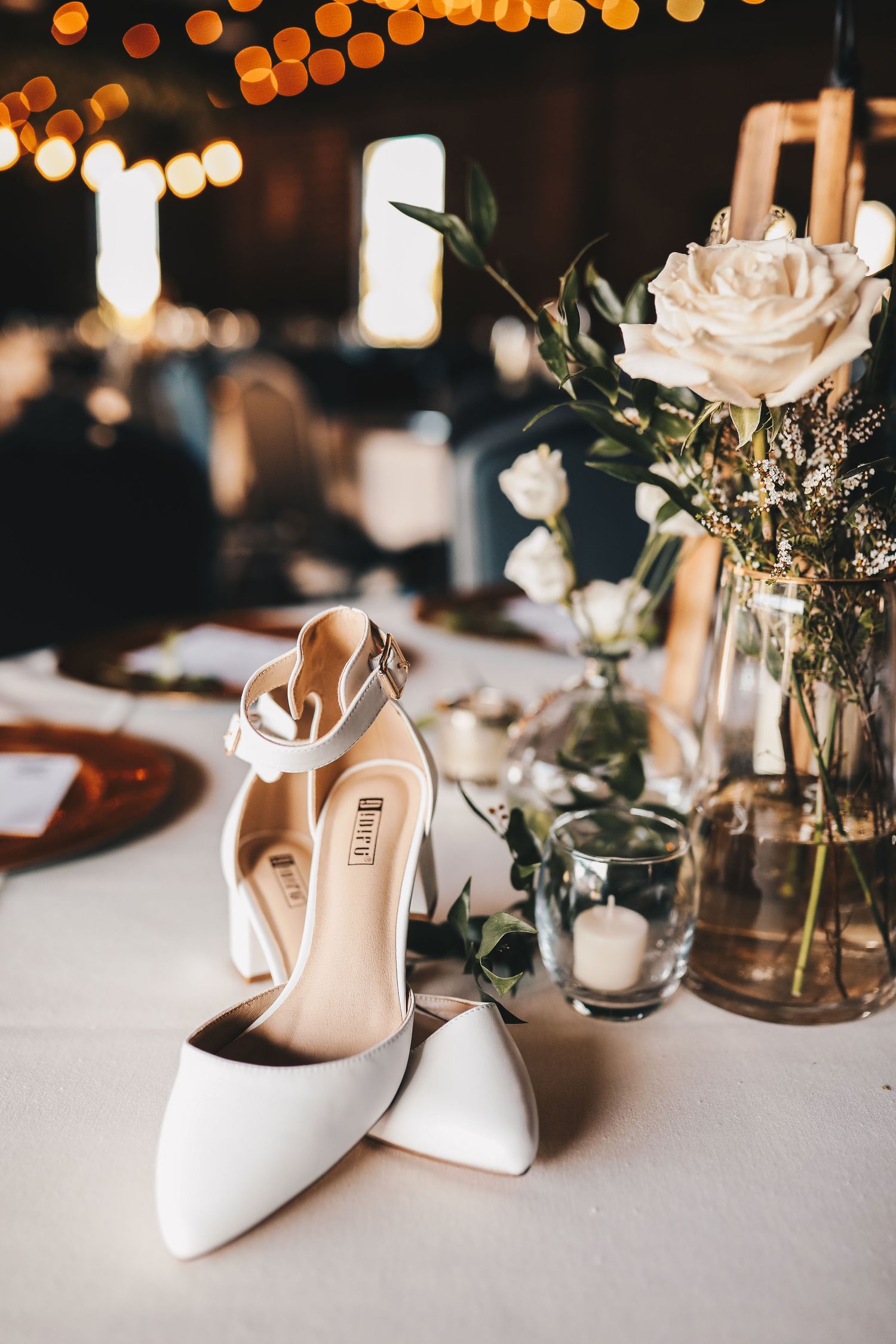 White bridal heels on reception table with white roses and candles in warm bokeh lighting.