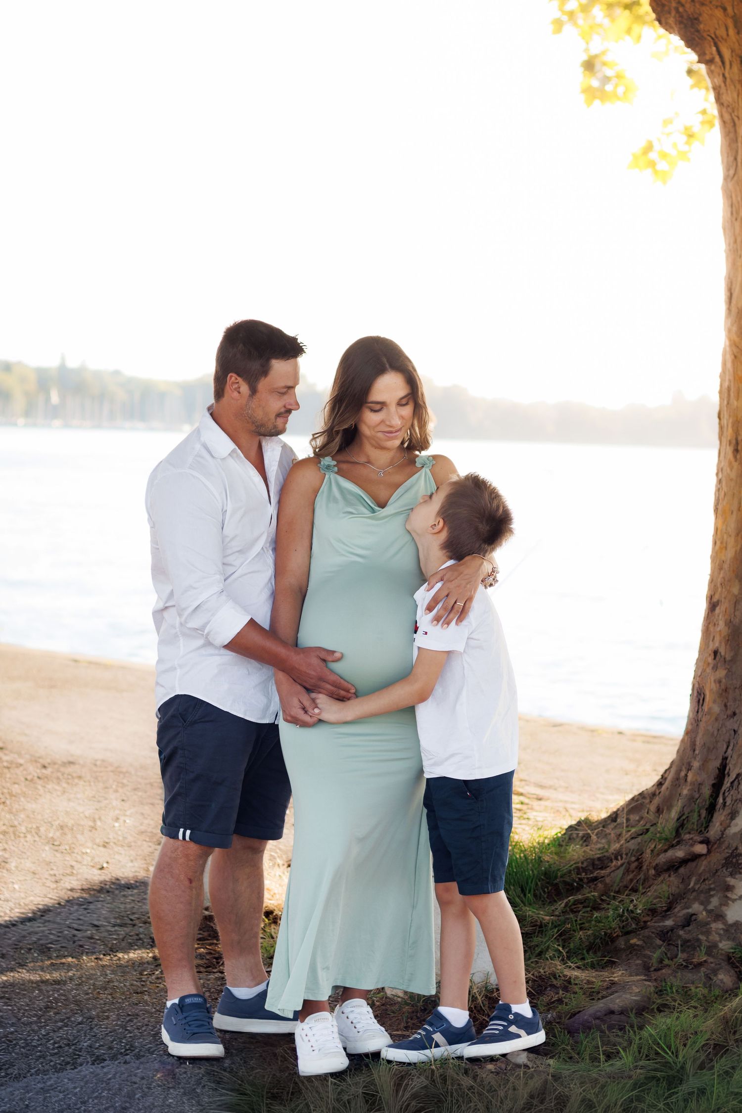 Séance photo grossesse en famille au bord de l’eau, un couple de futurs parents et leur fils aîné tendrement enlacés.