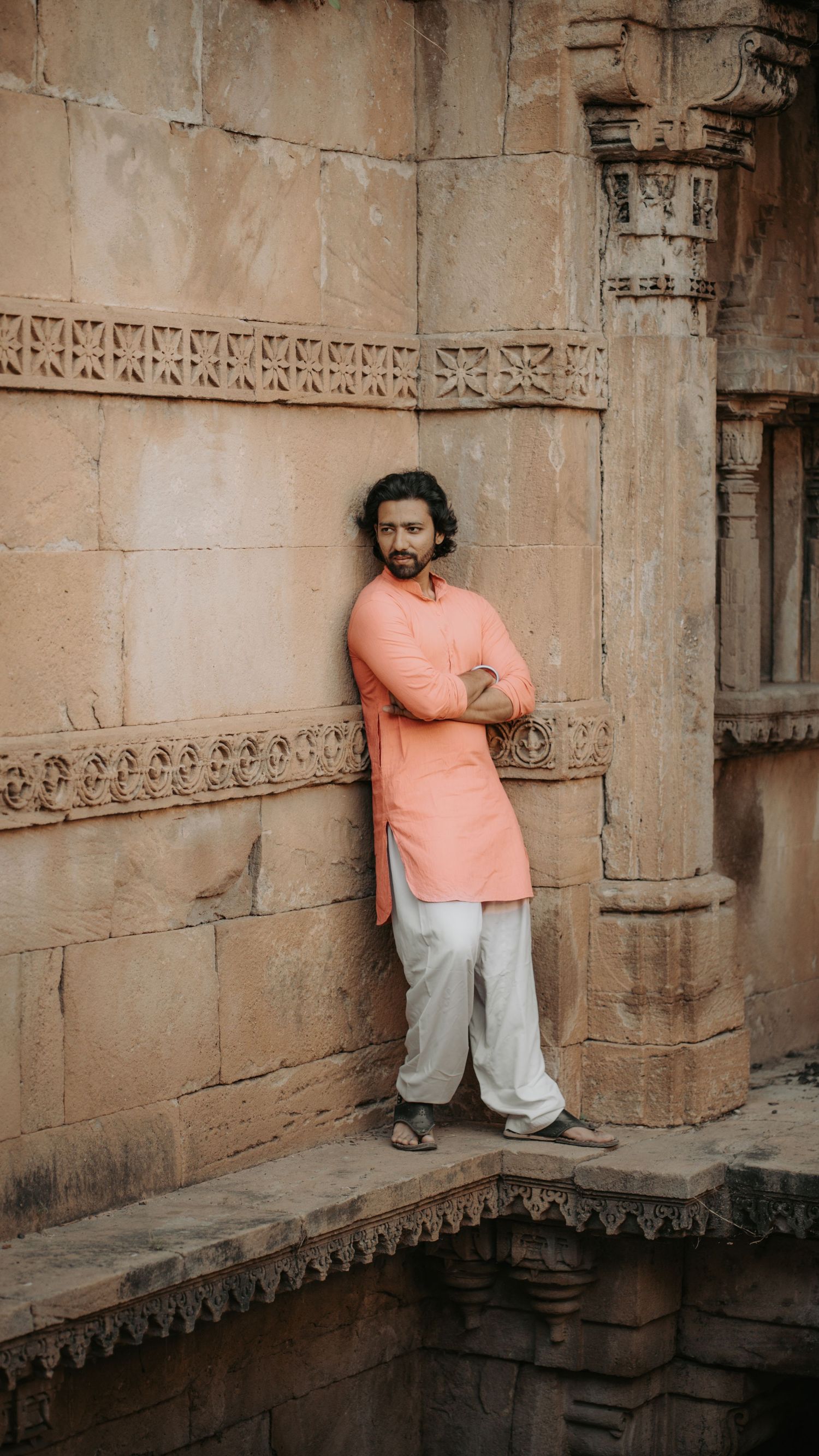 Man in peach kurta and white pants leaning against ancient stone temple wall with arms crossed.