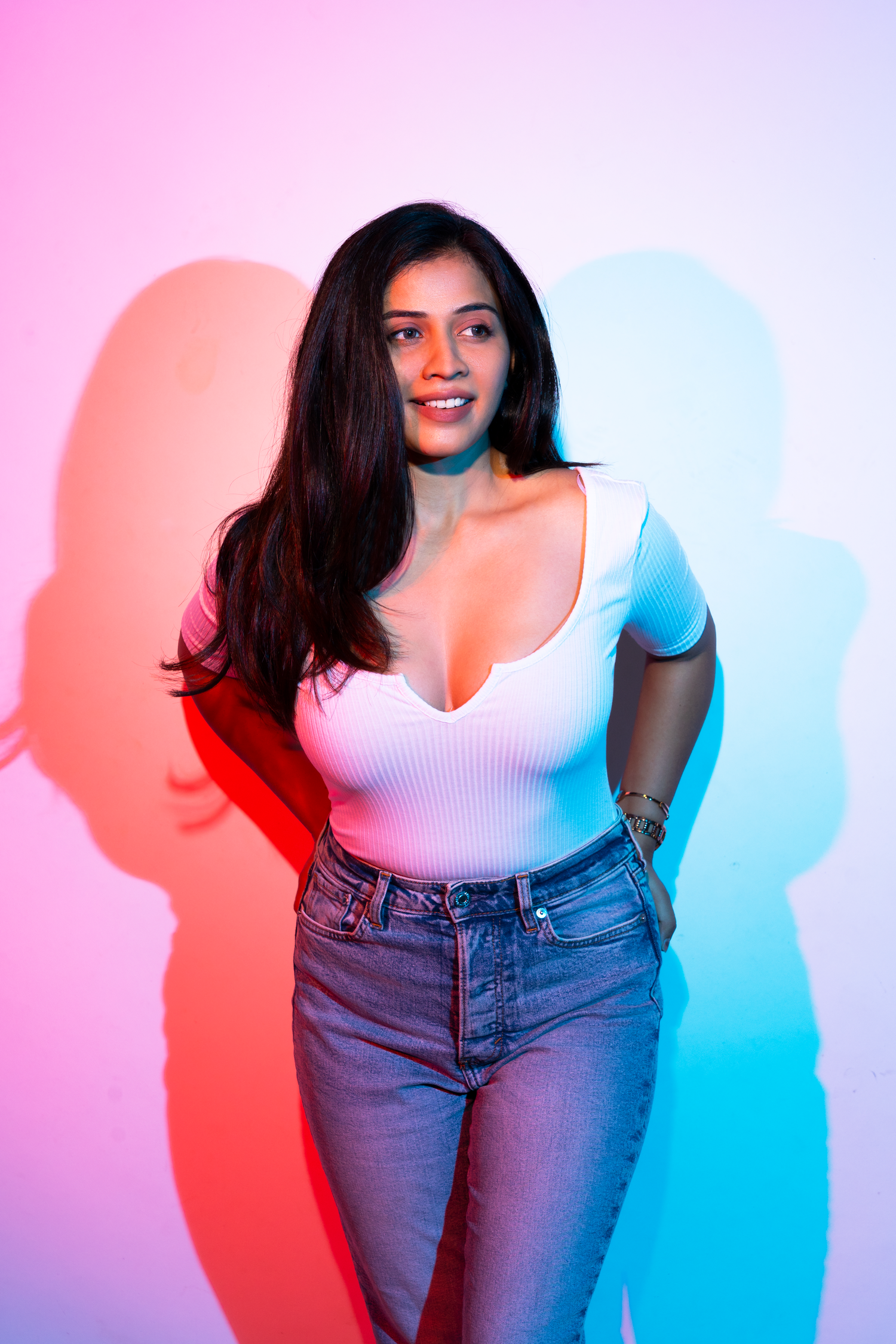 Young woman in white top and jeans posing confidently against a pink and blue neon-lit background.
