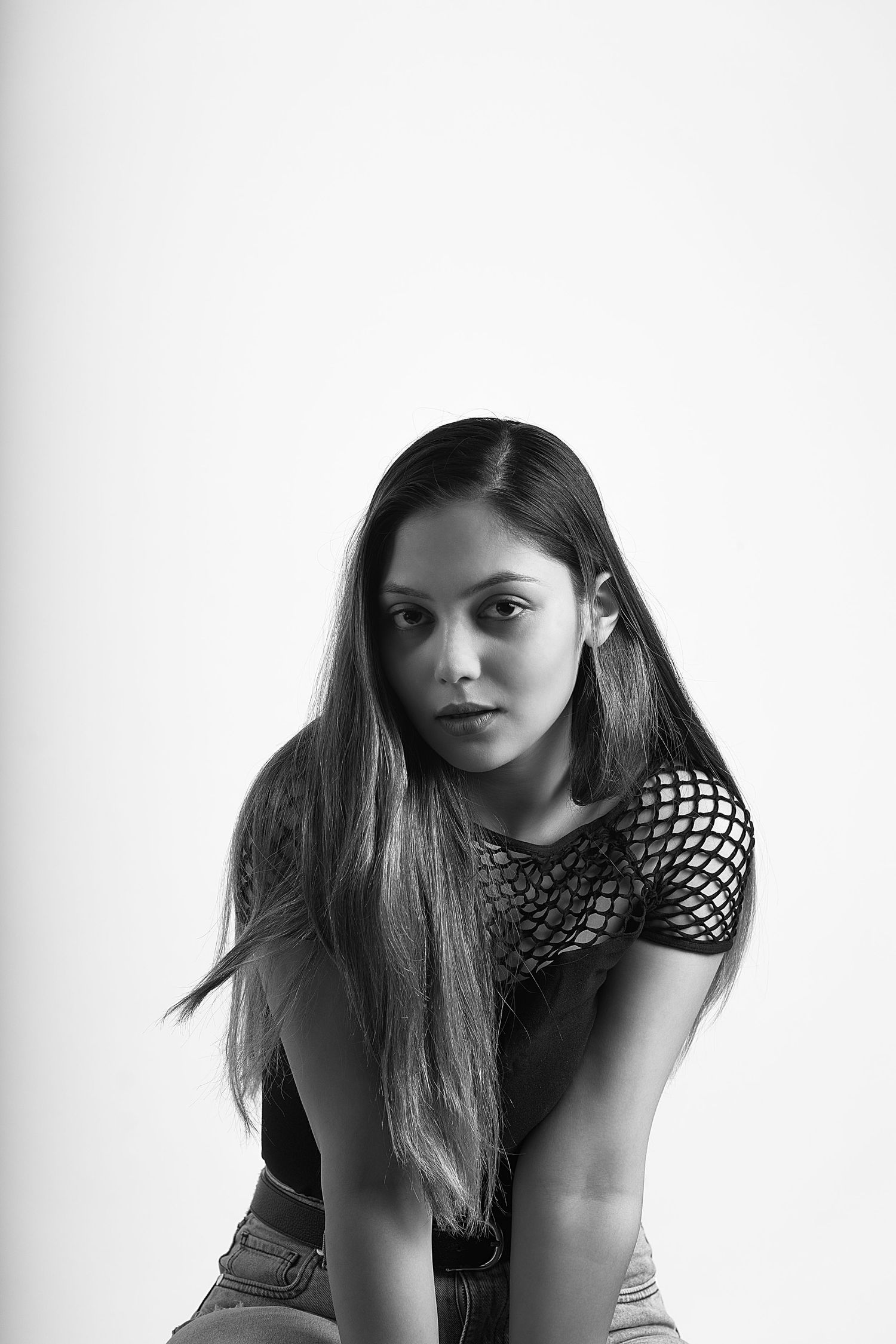 Black and white portrait of a young woman with long hair wearing a mesh top, leaning forward against white background.