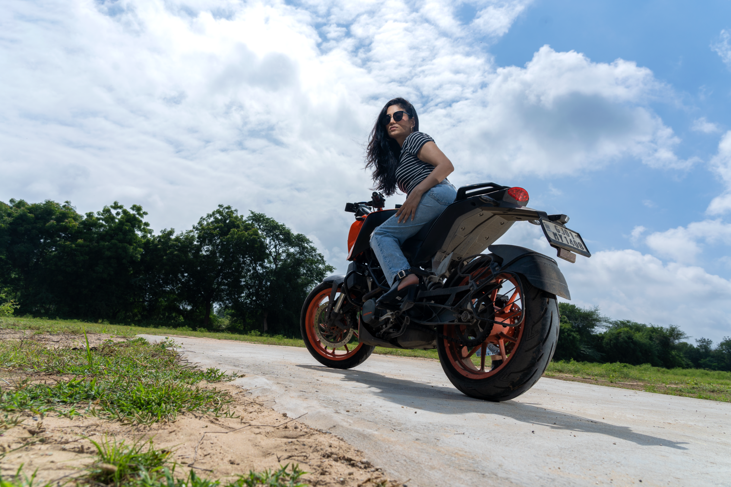 Woman in sunglasses riding an orange KTM motorcycle on a rural road under a blue cloudy sky.