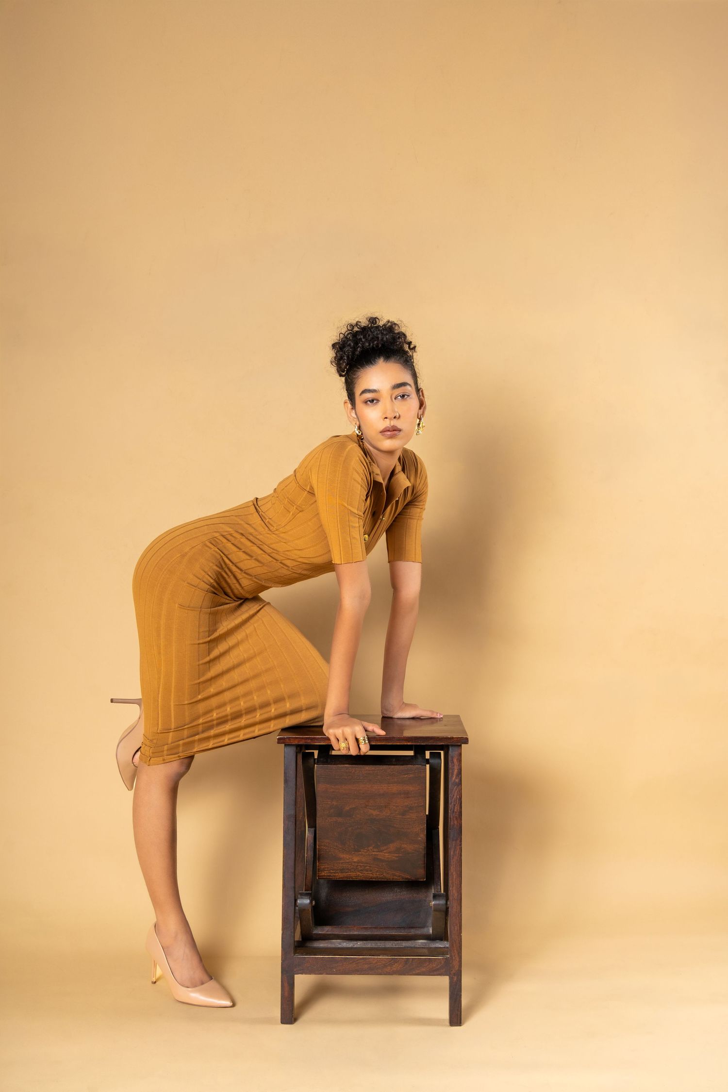 Woman in mustard midi dress leaning on wooden step stool against beige background.