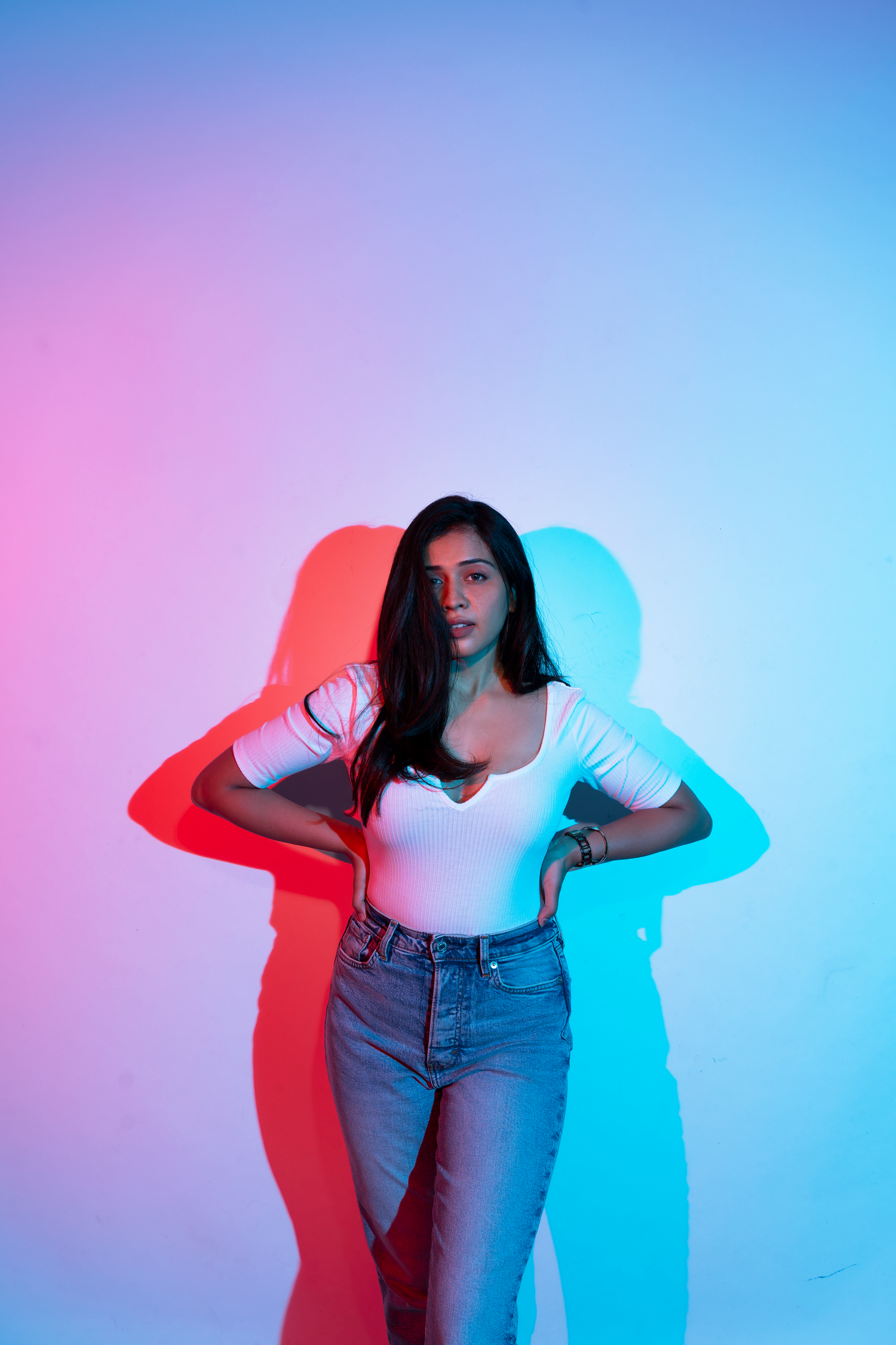 Young woman in white top and jeans poses confidently against a vibrant pink and blue neon lit studio background.