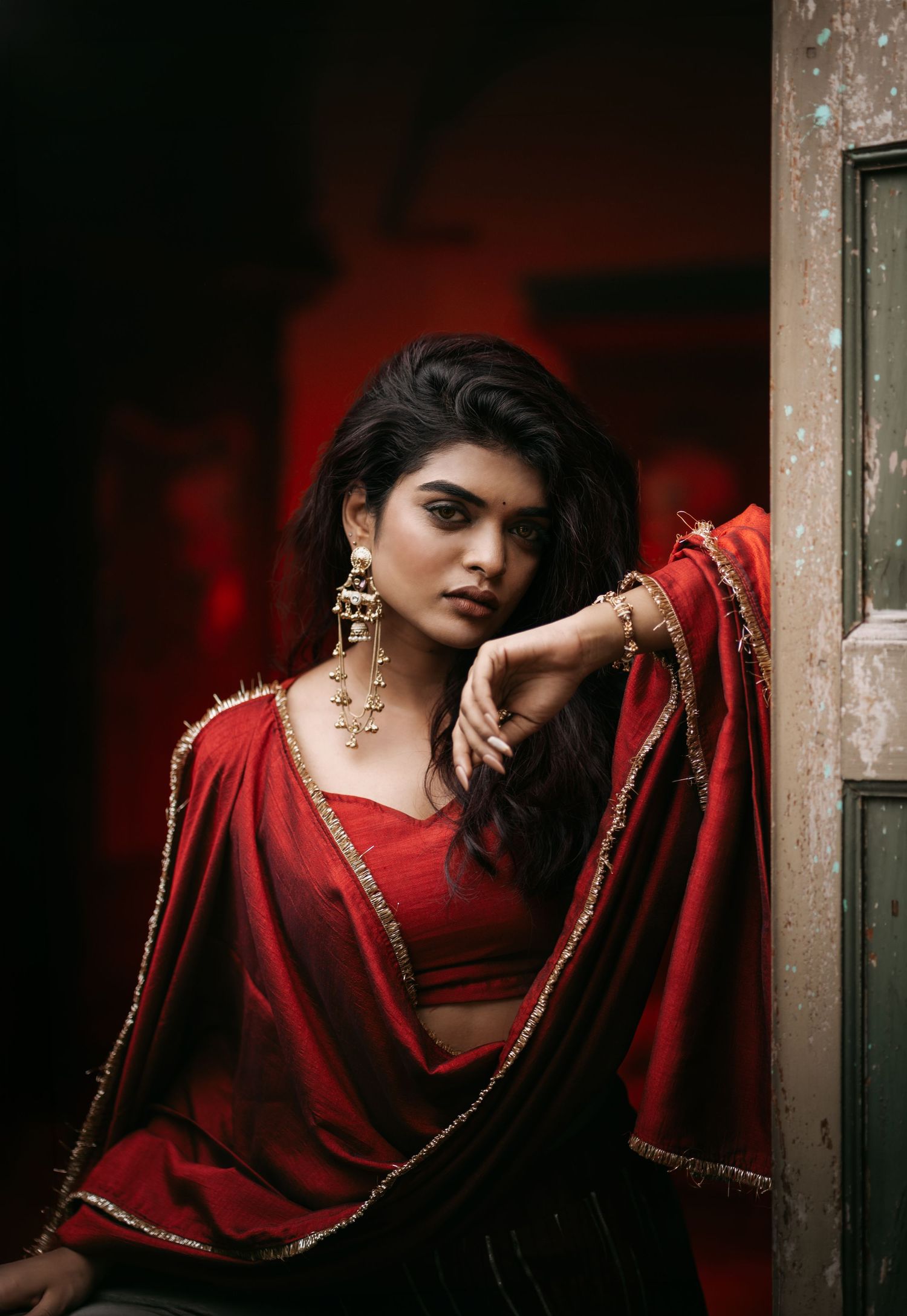 A young woman in a red saree with gold trim and jewelry poses elegantly against a weathered door in dramatic lighting.