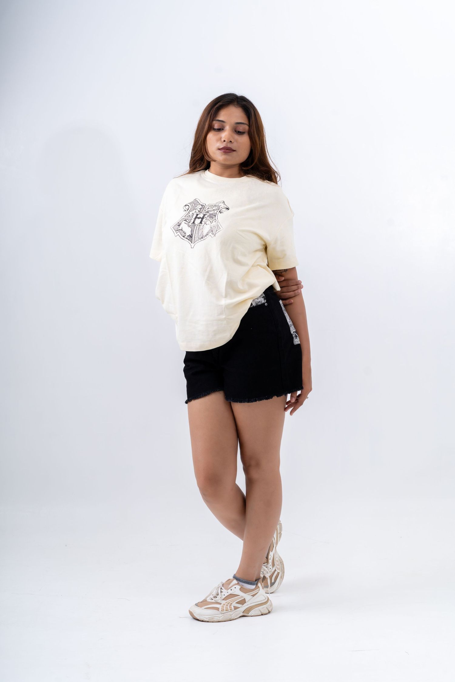 Woman wearing cream Harry Potter graphic tee, black denim shorts, and white sneakers posing against white background.