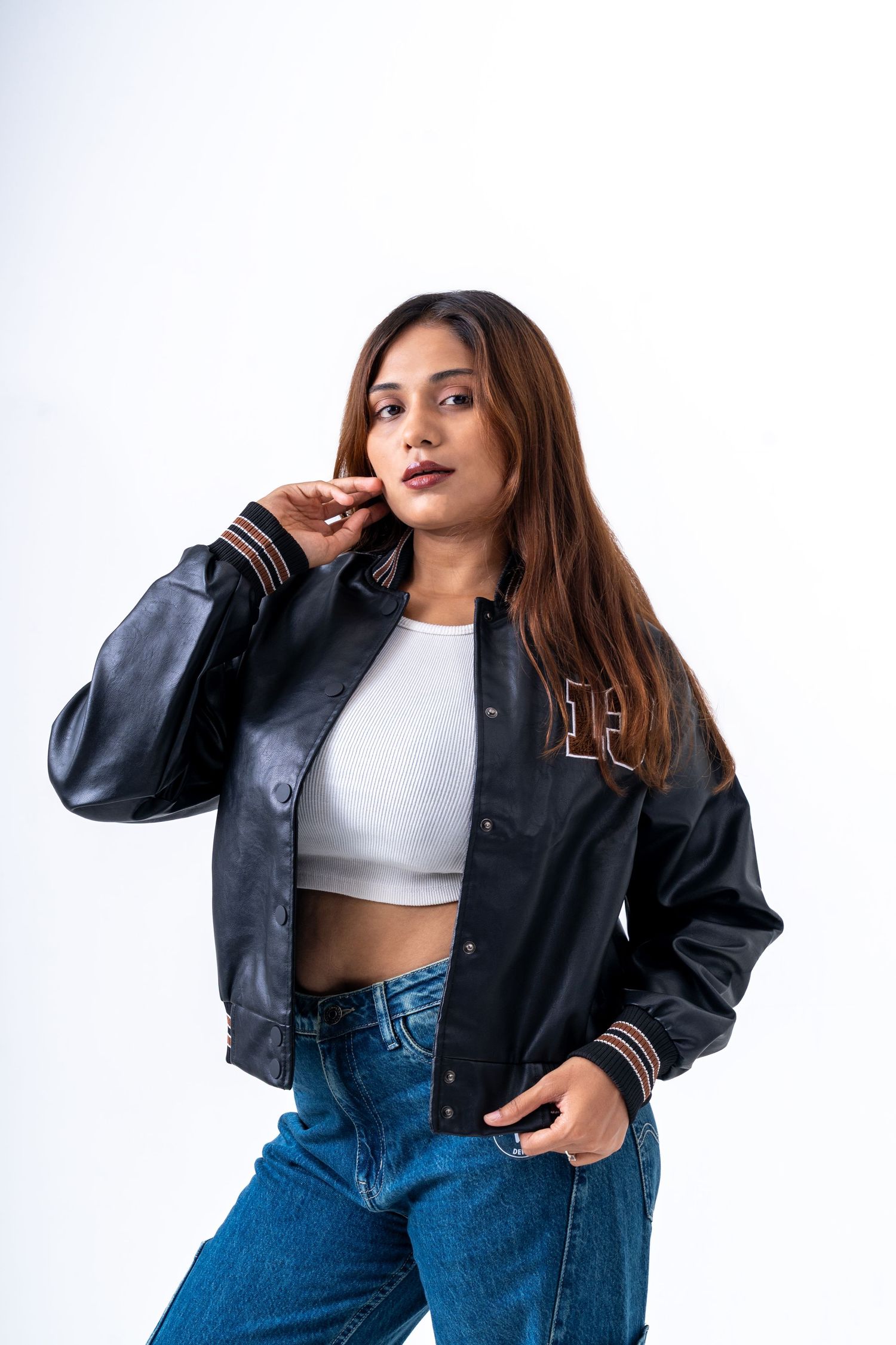 Young woman in black leather varsity jacket, white crop top, and blue jeans posing confidently on white background.