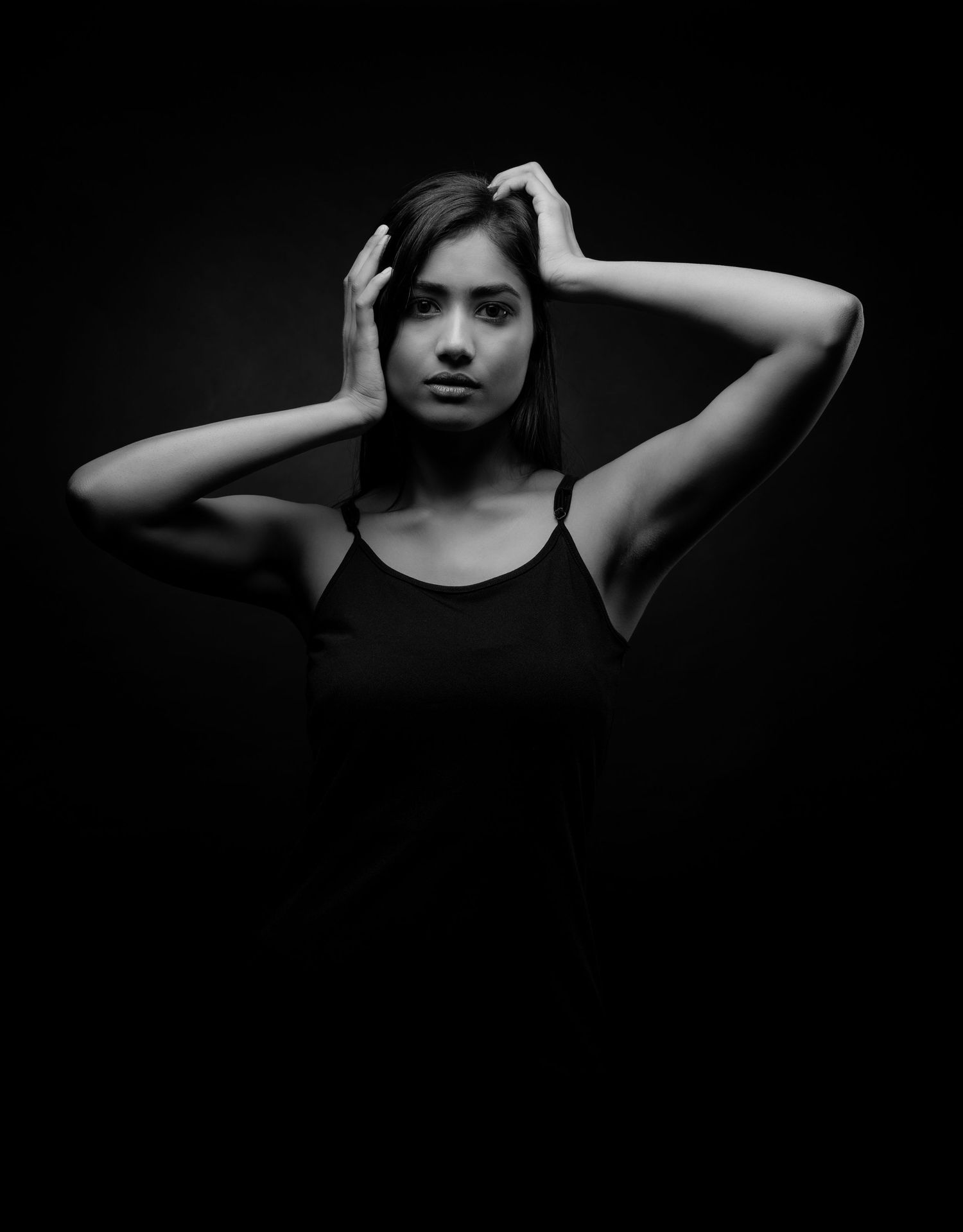 Black and white portrait of a young woman in a black tank top posing with hands on her head against dark background.
