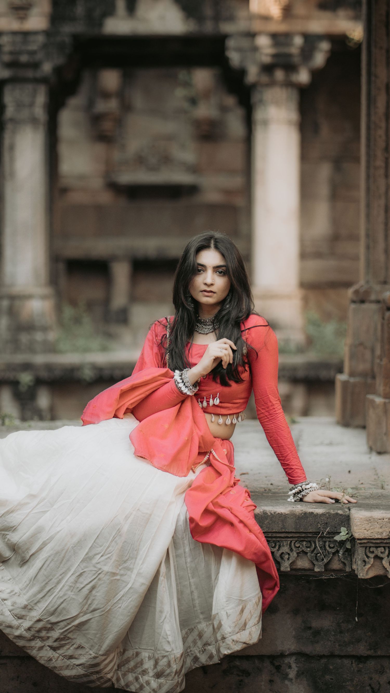 Young woman in red and white traditional Indian lehenga sits gracefully on ancient temple steps, adorned with silver jewelry.