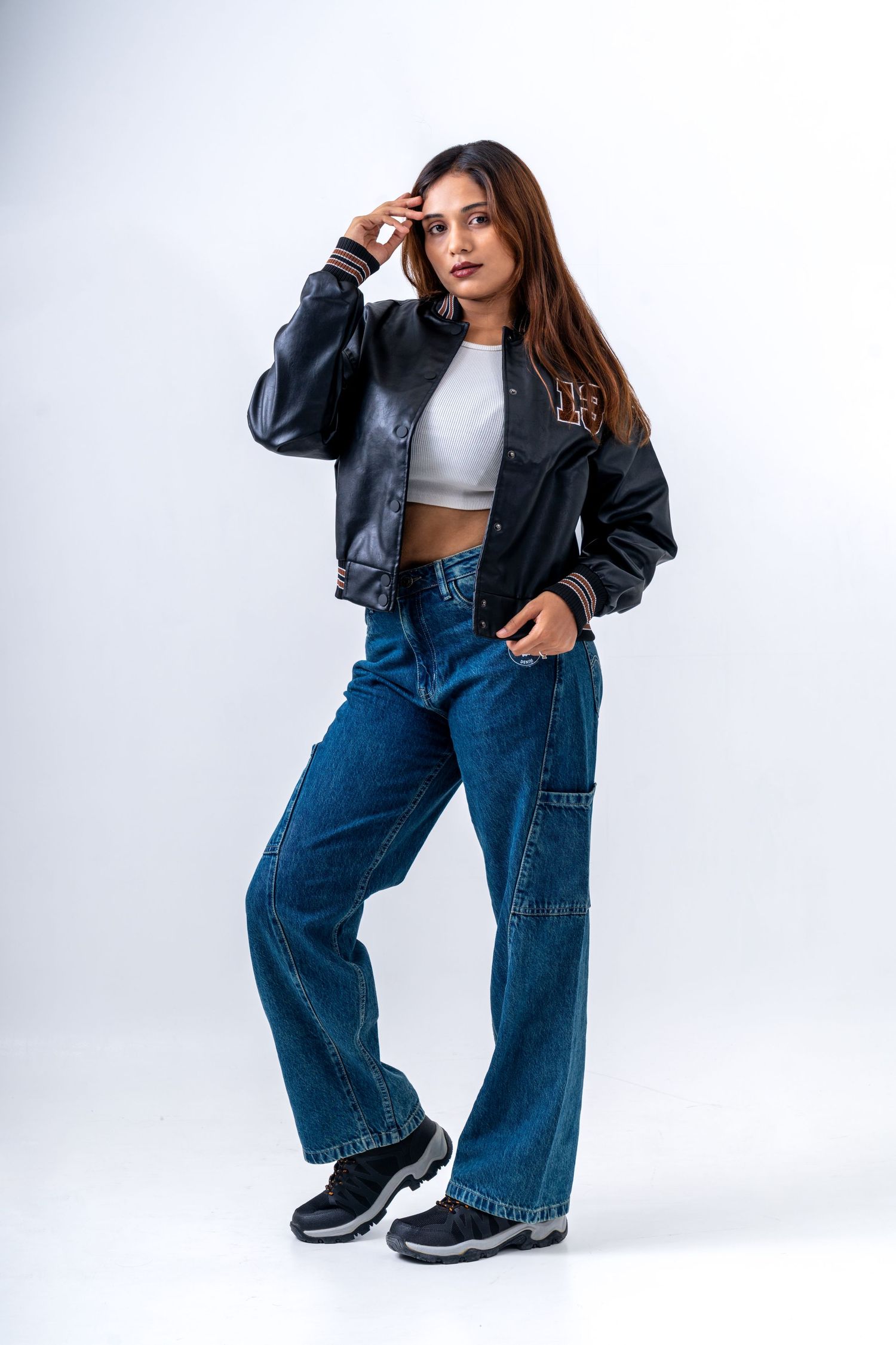 Young woman in black leather bomber jacket, white crop top, wide-leg blue jeans, and sneakers posing on white background.