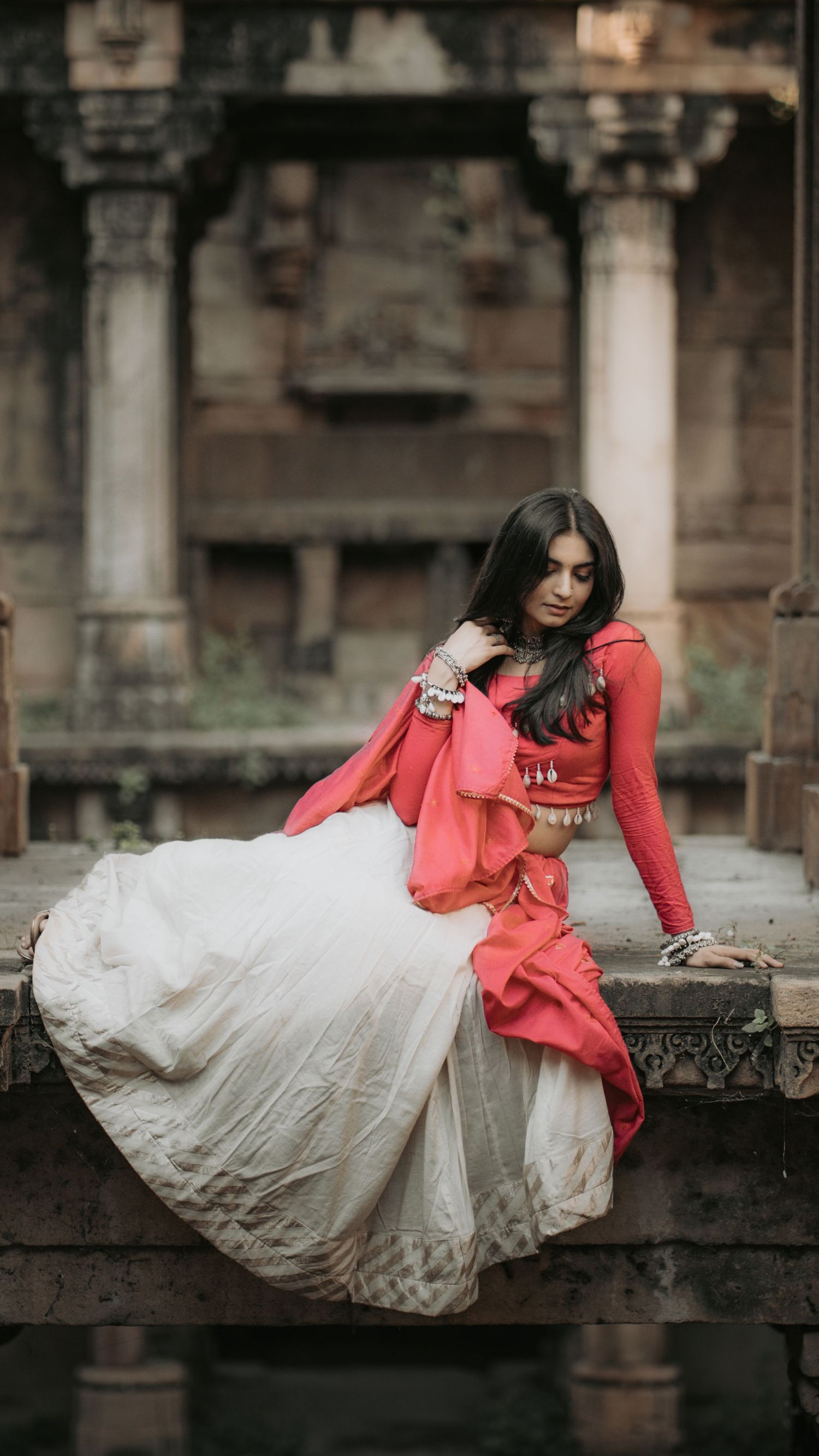 A woman in a red top and white lehenga skirt sits gracefully on stone steps at an ancient Indian temple.