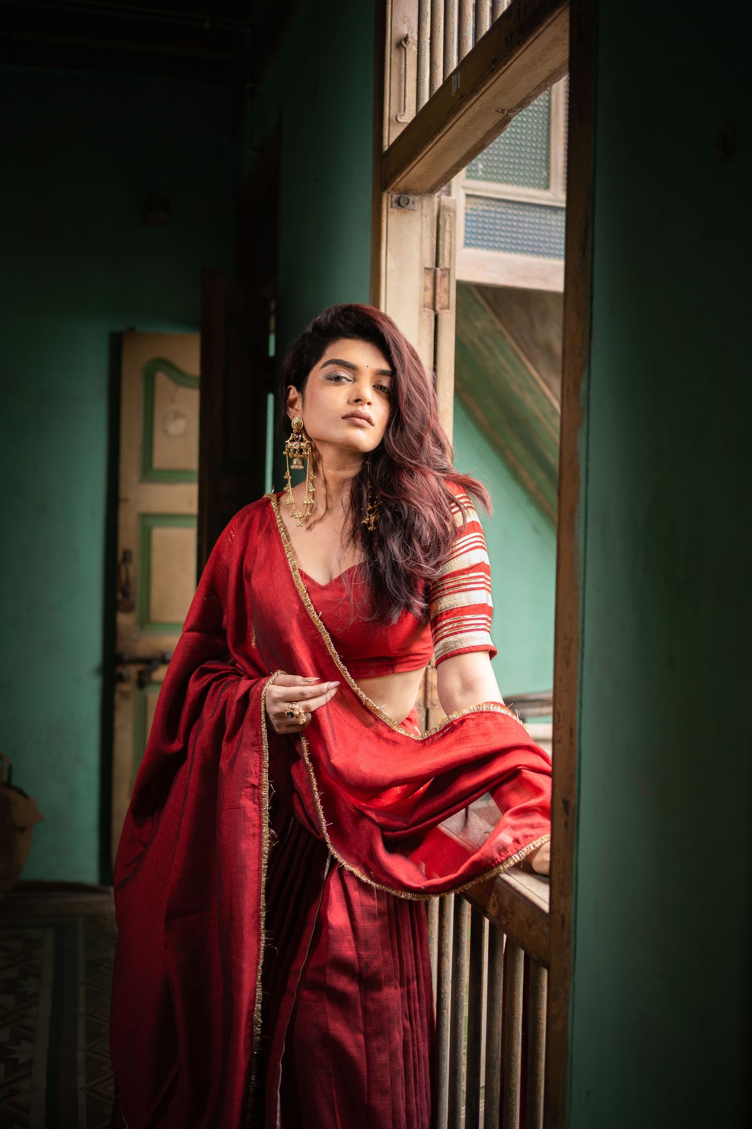 A young woman in a red saree with gold trim poses gracefully by a vintage window in an elegant interior.
