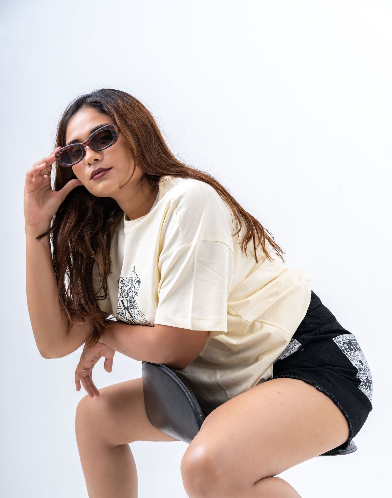 Woman posing in cream graphic tee, black shorts, and brown sunglasses against white background.