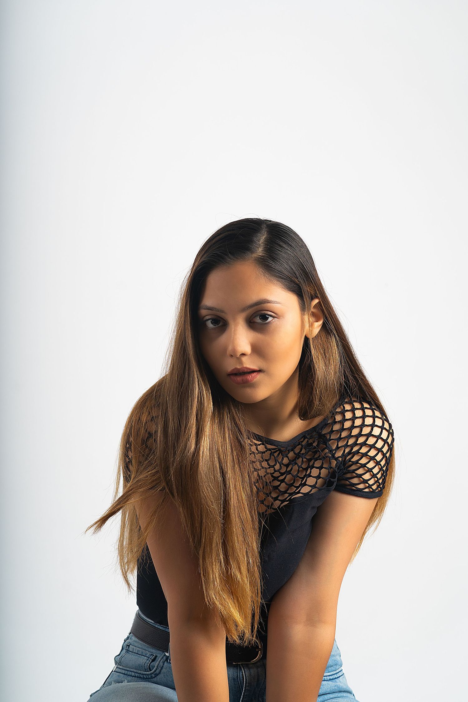Young woman with long hair wearing a black mesh top and jeans, posing confidently against a white background.