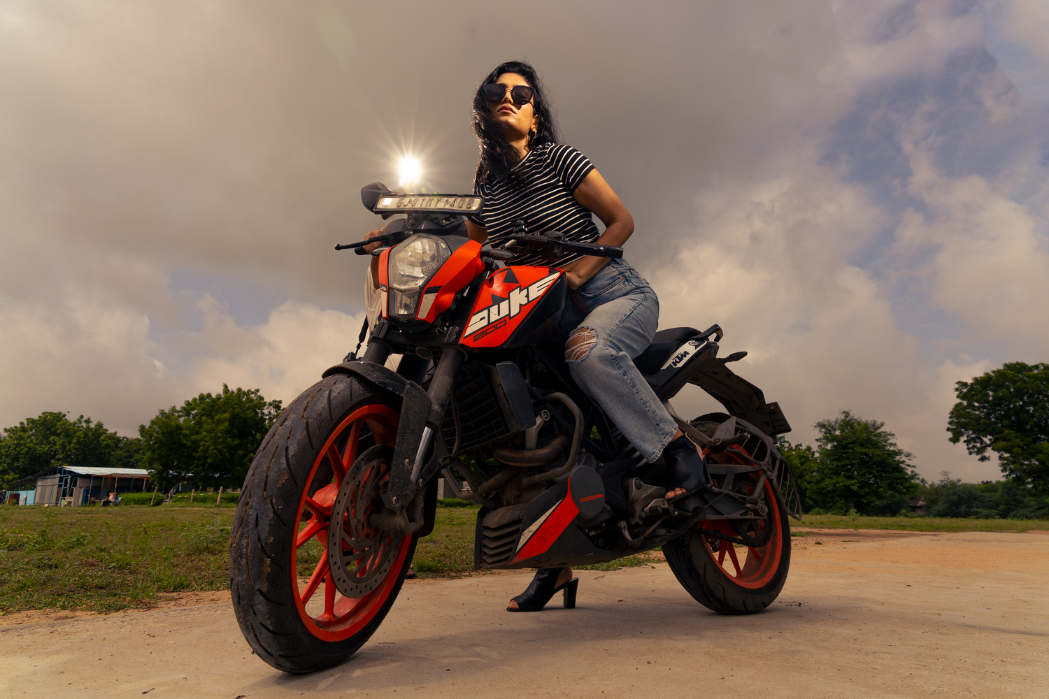 Woman in jeans and striped top posing on a red and black motorcycle outdoors under a dramatic cloudy sky.
