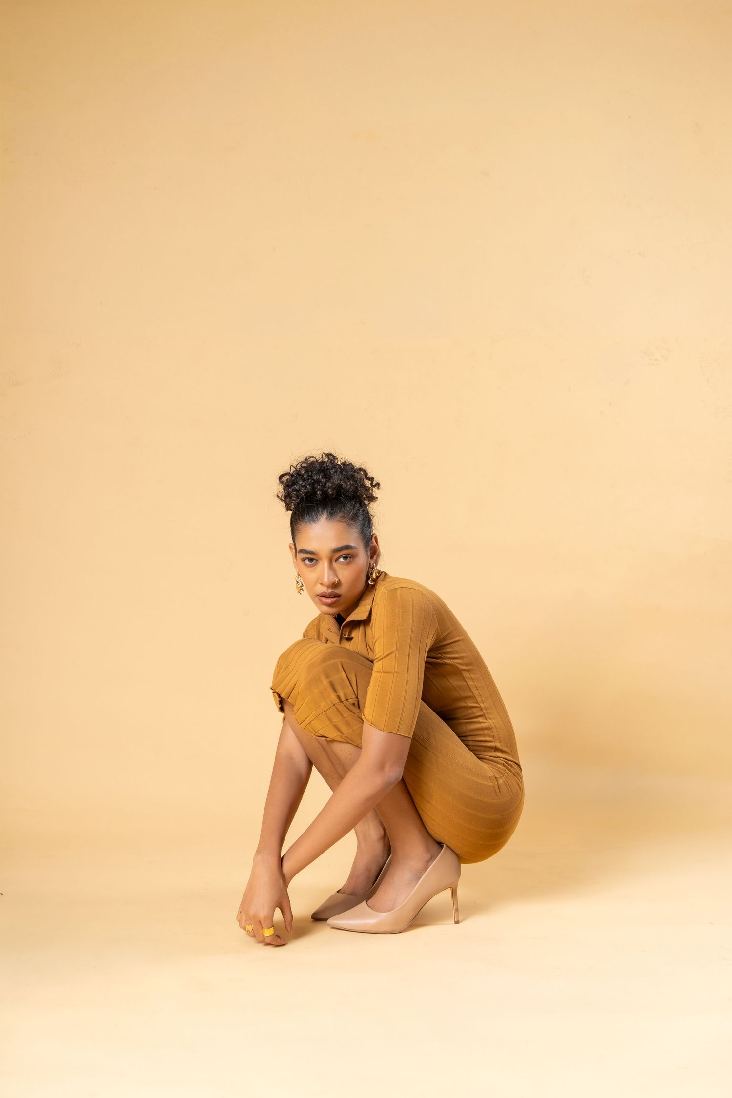 A young woman in a mustard yellow dress and nude heels crouches gracefully against a beige background.