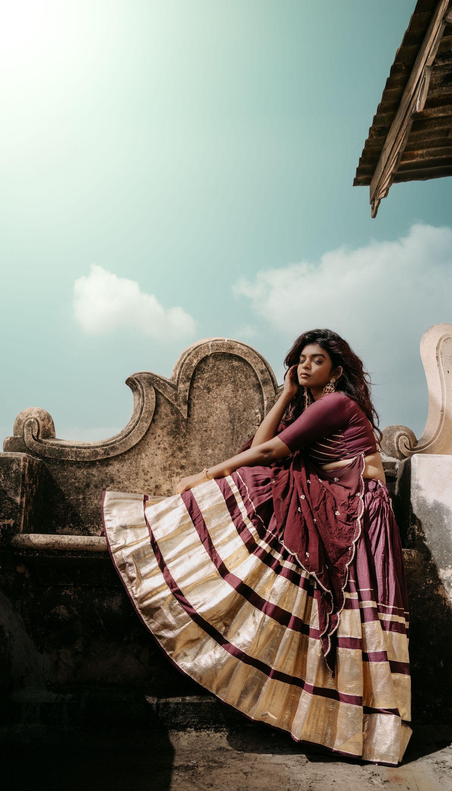 A woman in a flowing maroon and gold striped lehenga poses gracefully on an ancient stone structure under a blue sky.