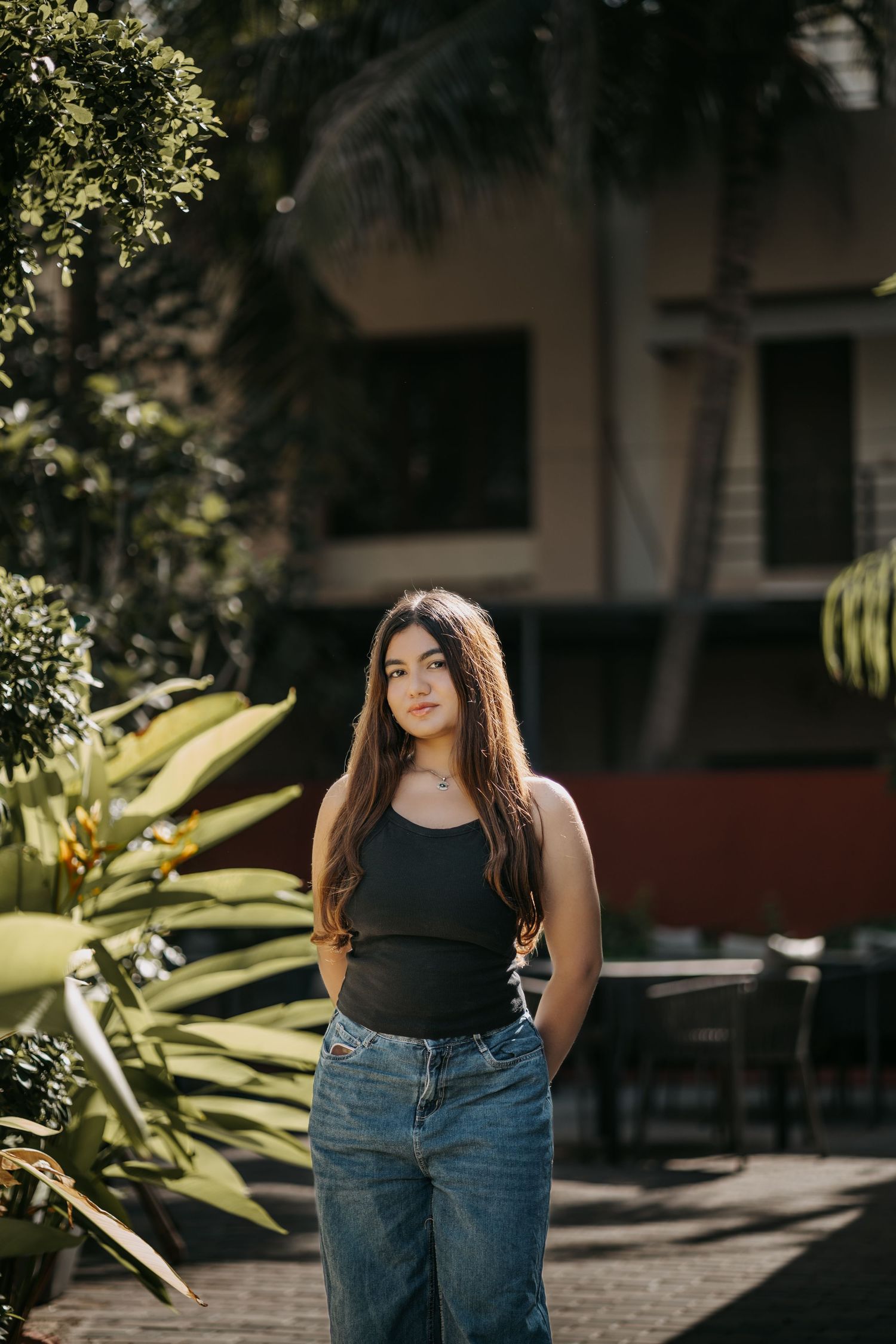 Young woman in black tank top and blue jeans poses outdoors among tropical plants near a building.