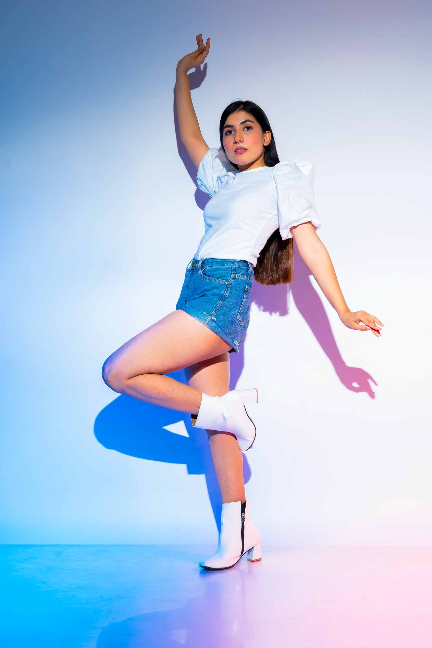 Young woman in white puffed sleeve top, denim mini skirt, and white boots poses dynamically under blue and pink studio lights.