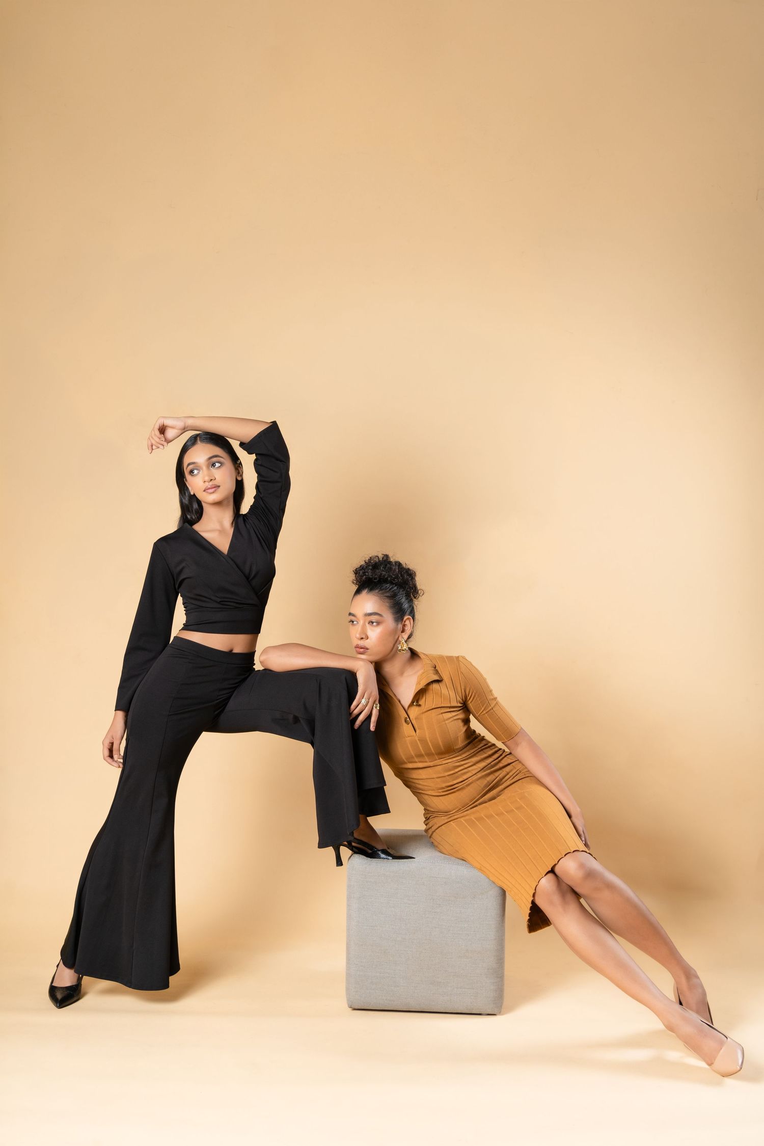 Two models pose elegantly, one in black wide-leg pants and crop top, another in tan dress on a cube, against beige backdrop.