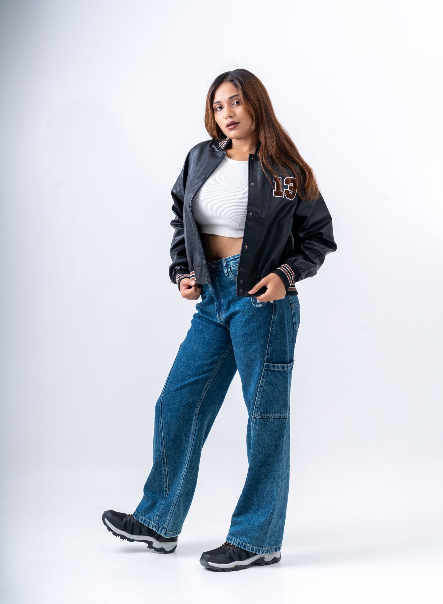 Woman in black varsity jacket, white crop top, wide-leg blue jeans, and sneakers posing on white background.