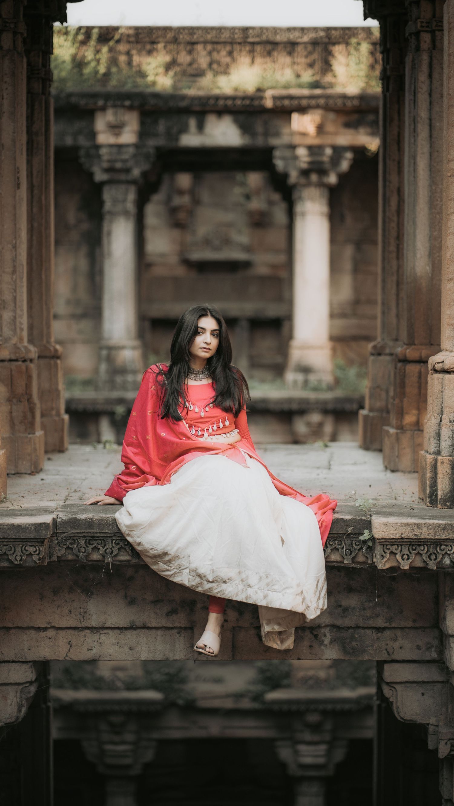 Woman in red dupatta and white lehenga sitting gracefully at an ancient Indian temple with ornate stone columns.