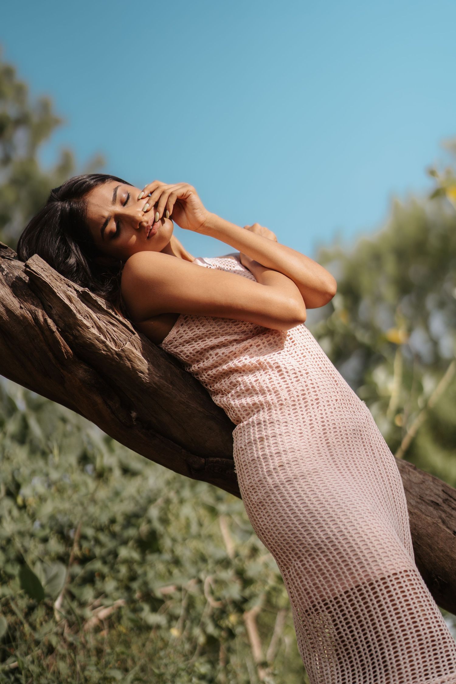 Woman in pink crochet dress leaning against a large log outdoors with blue sky and green foliage background.