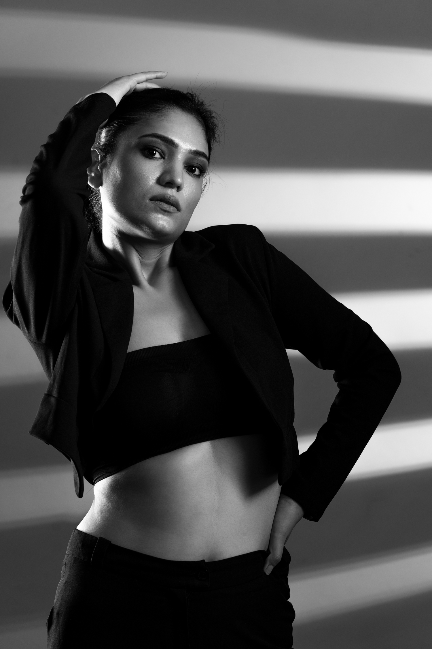 Black and white fashion portrait of a woman in crop top and blazer posing dramatically against striped light background.