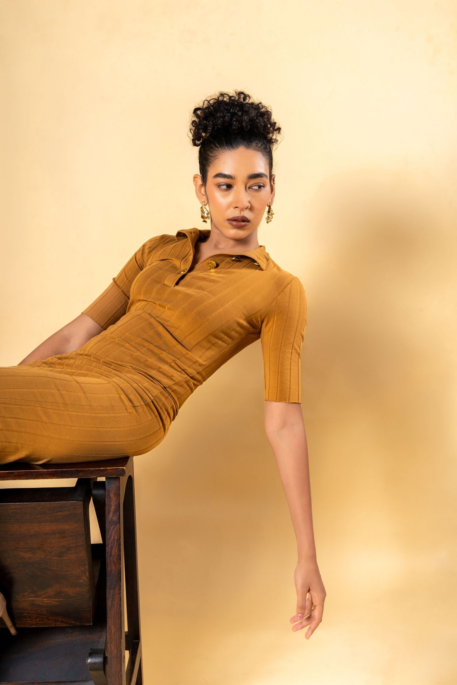 A woman in a mustard yellow fitted dress poses elegantly on a wooden chair against a warm beige background.