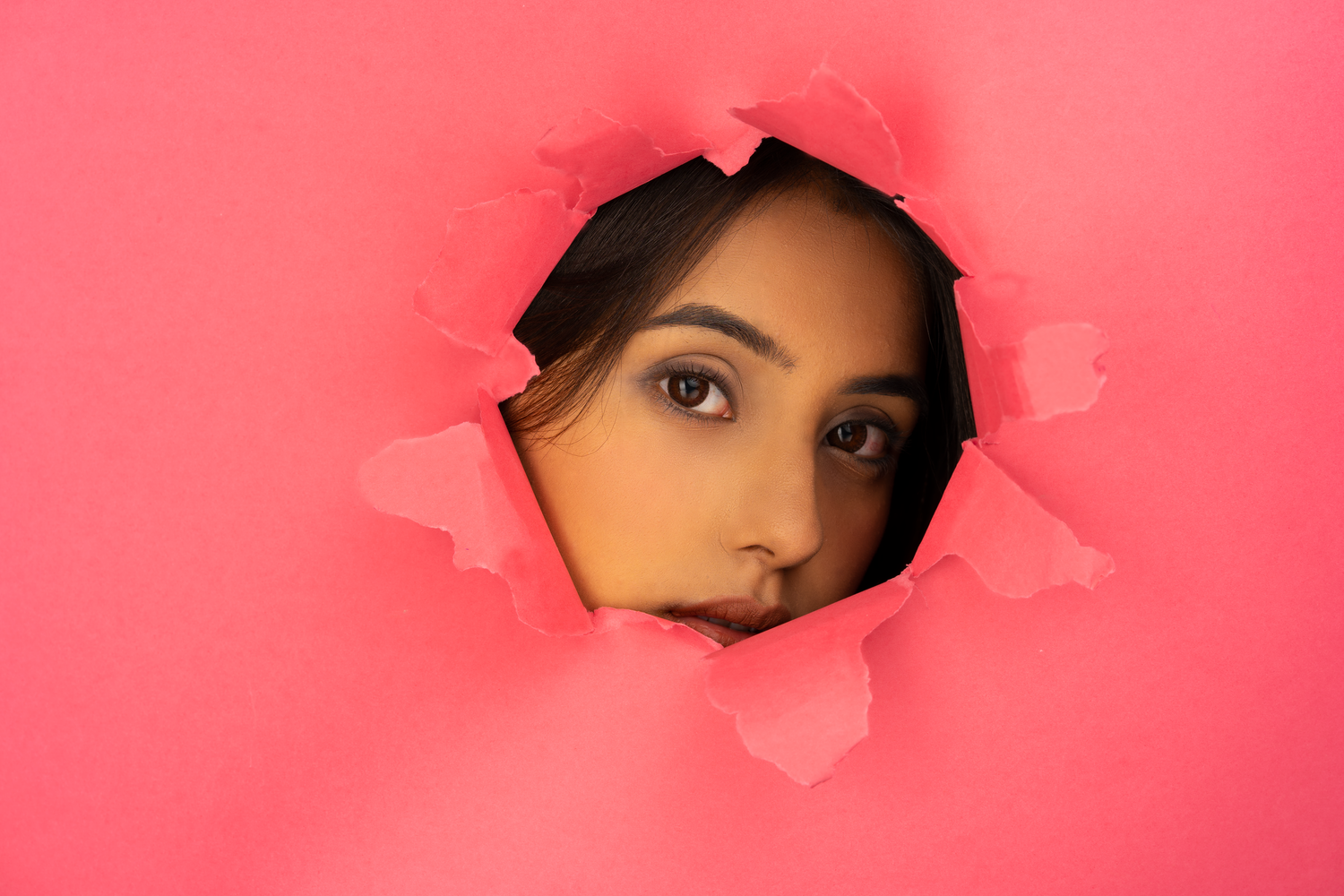 A young woman peers through a torn hole in a bright pink paper background with a curious expression.