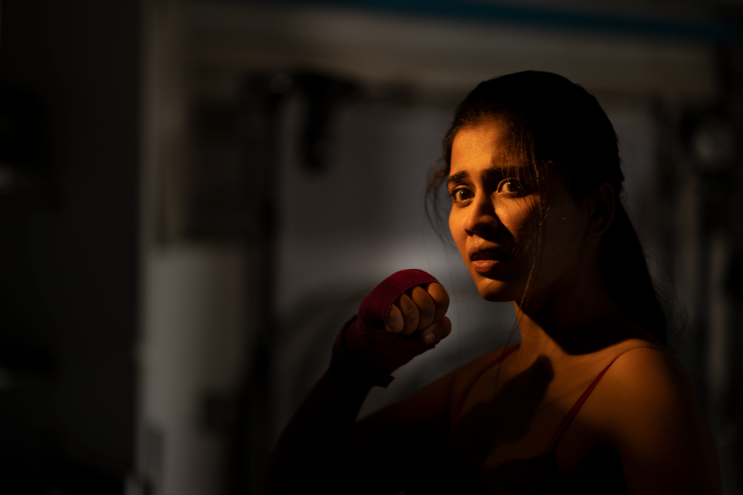 A focused female boxer with red hand wraps trains in a dimly lit gym, poised in a fighting stance.