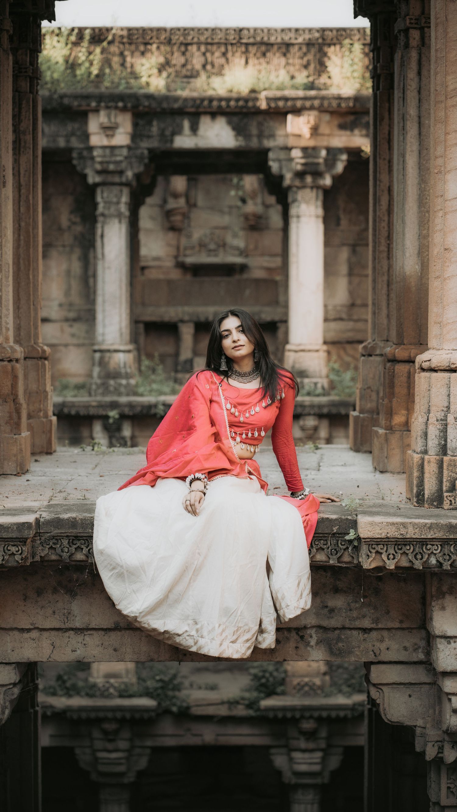 Woman in red and white traditional Indian attire sitting gracefully at an ancient stone temple architecture.