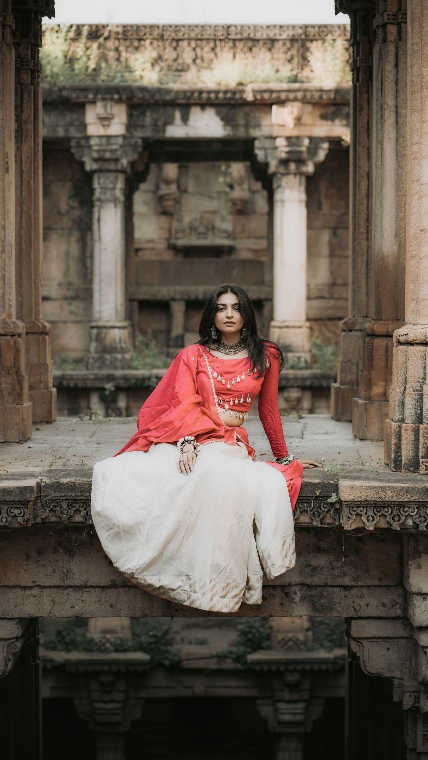 Woman in red and white traditional Indian lehenga sitting at ancient stone temple ruins.