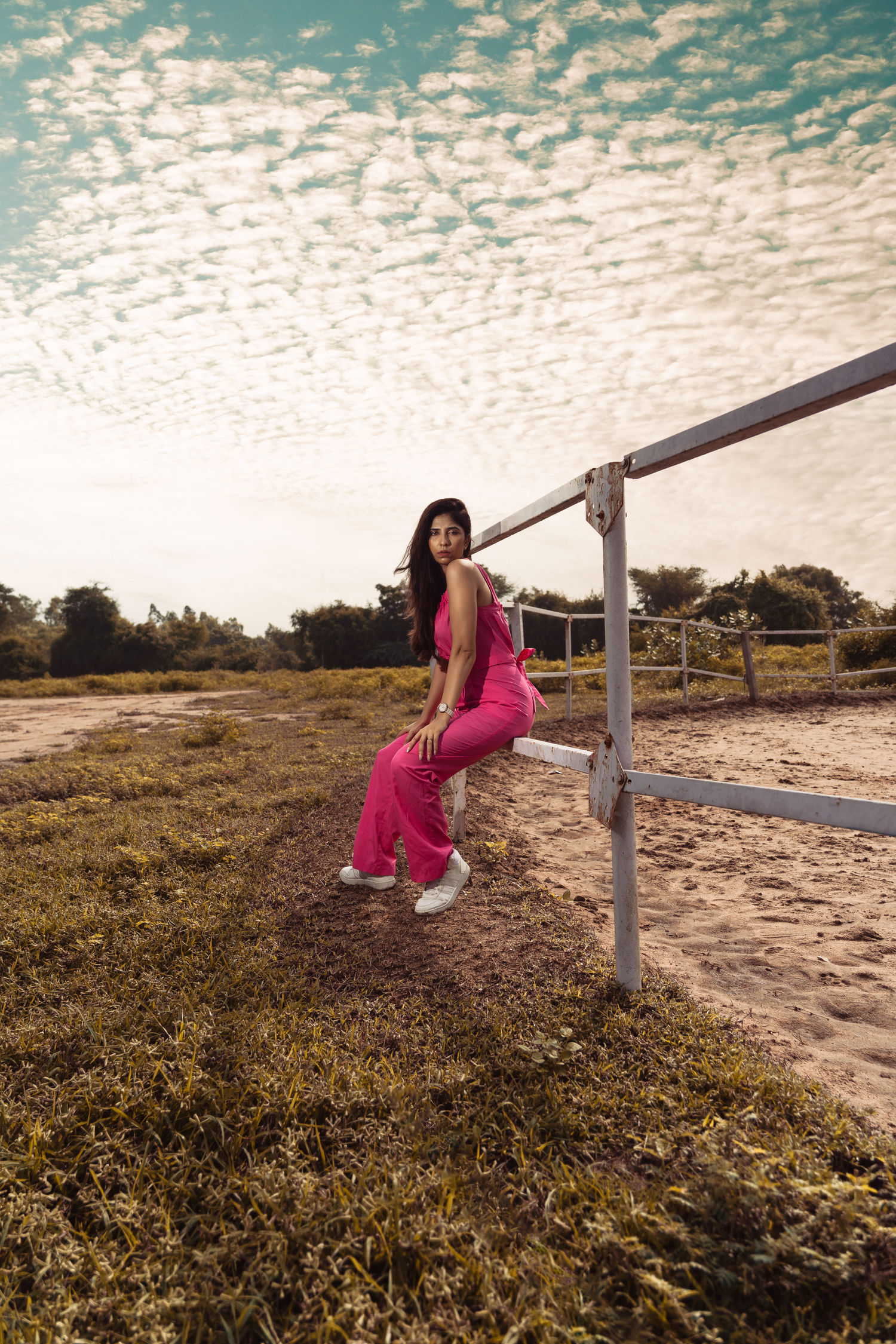Woman in pink jumpsuit sitting on white fence rail in open field under dramatic cloudy sky.